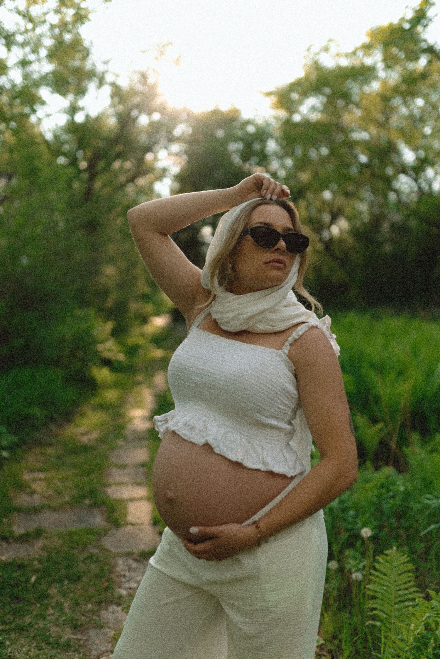 Pregnant woman in white crop top and pants poses outdoors in sunglasses, cradling her baby bump in a lush garden.
