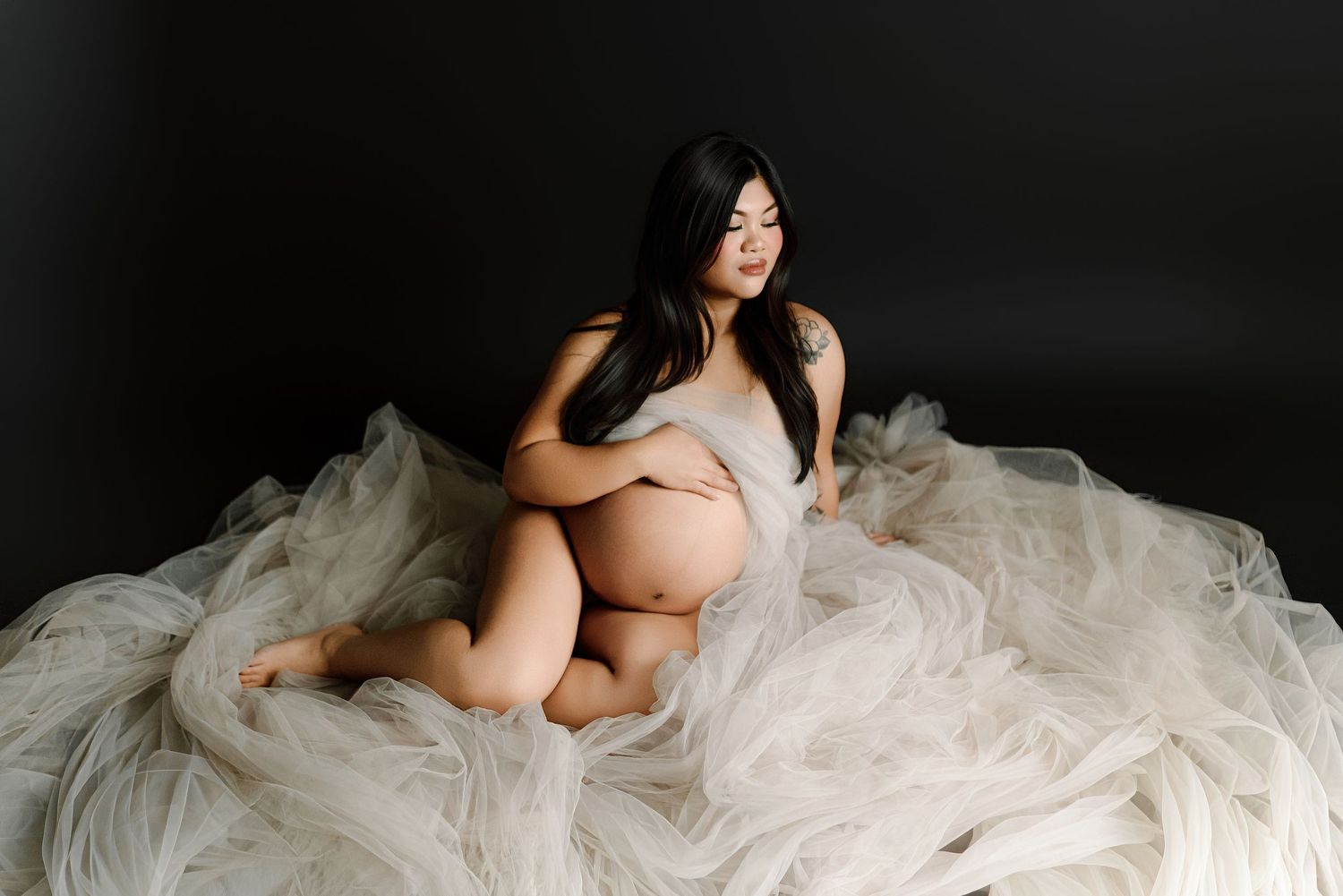 Pregnant woman in sheer white tulle fabric sitting gracefully against a dark black studio background.