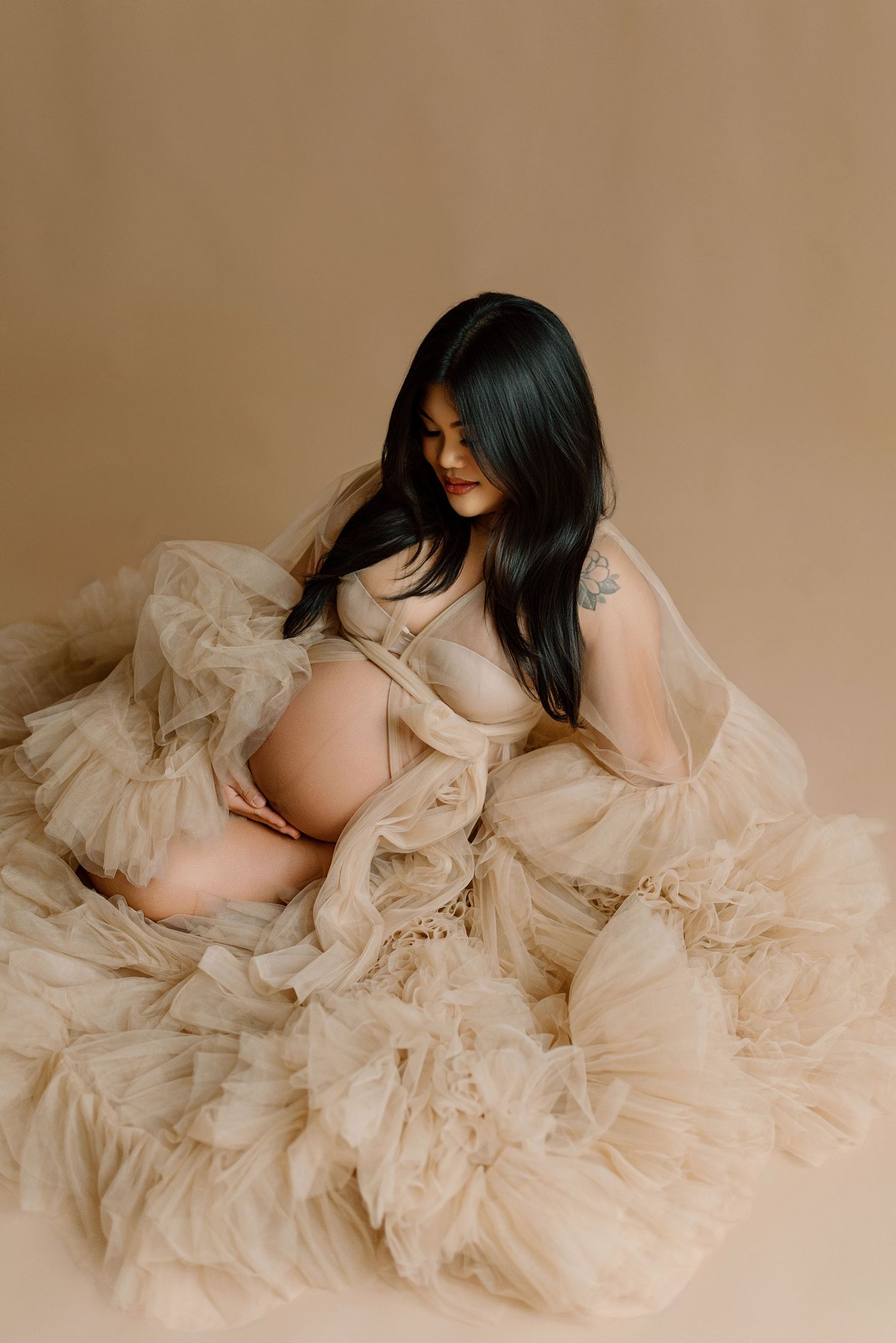Pregnant woman in flowing beige tulle robe sitting gracefully against neutral studio background.