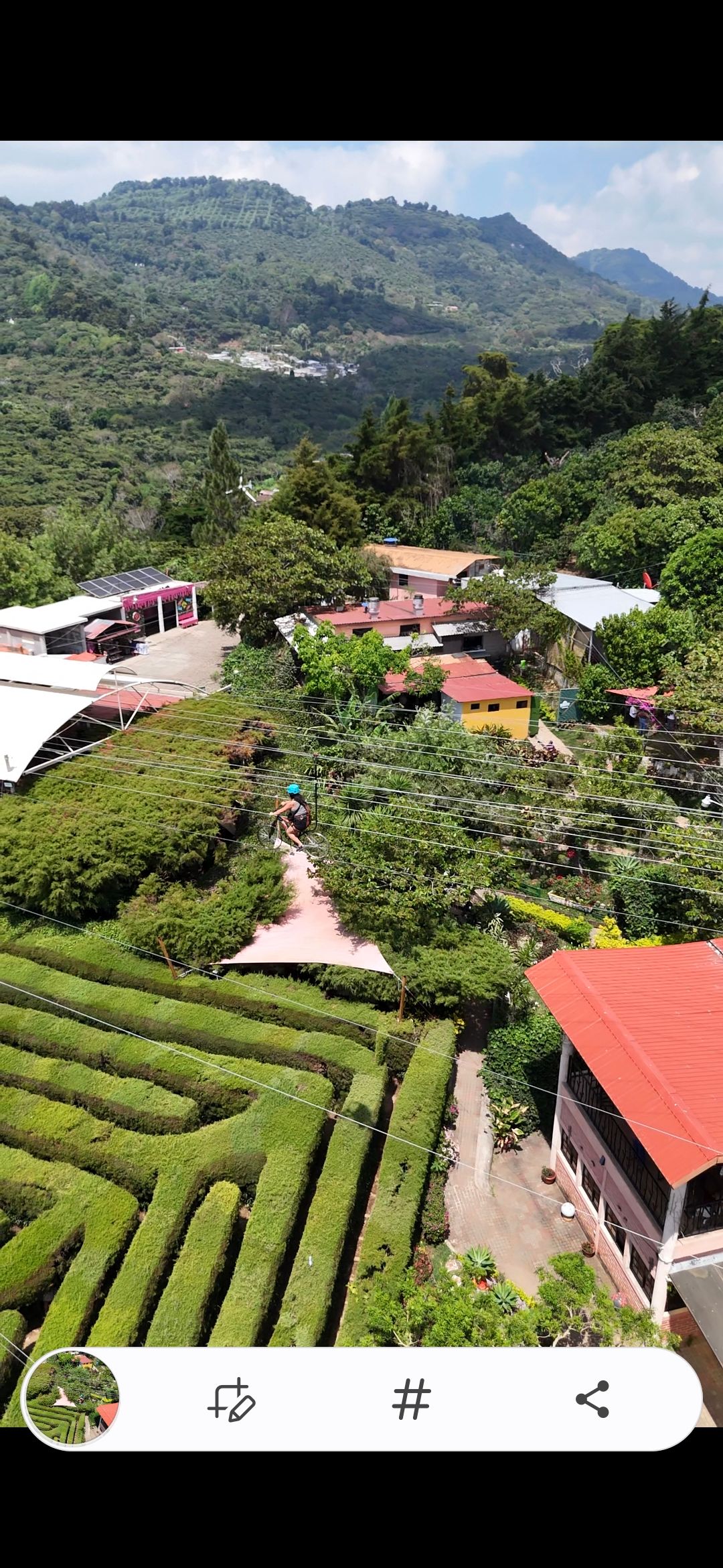 Aerial view of a lush green hedge maze surrounded by colorful buildings and mountain scenery.