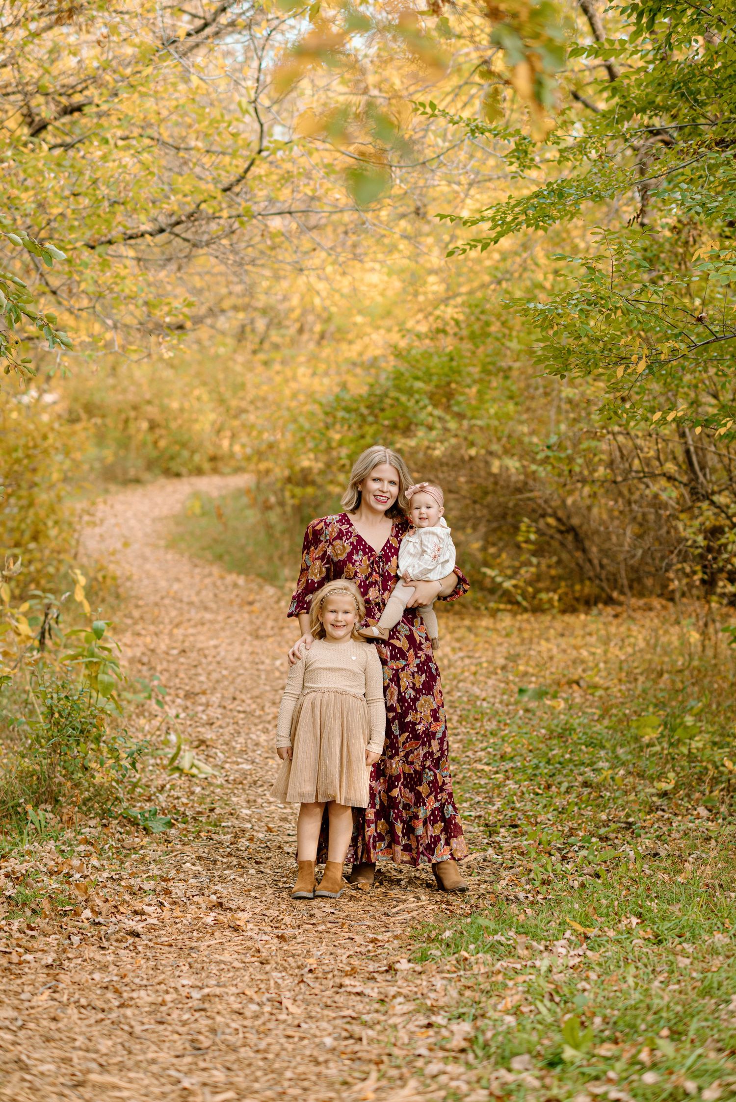 A mother in a floral dress holds her baby while embracing her young daughter on a leaf-covered autumn path.