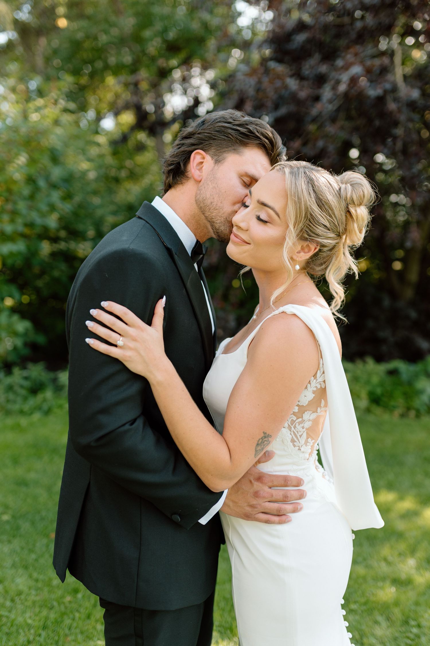 Groom tenderly kissing bride on forehead while embracing her in a garden wedding portrait.
