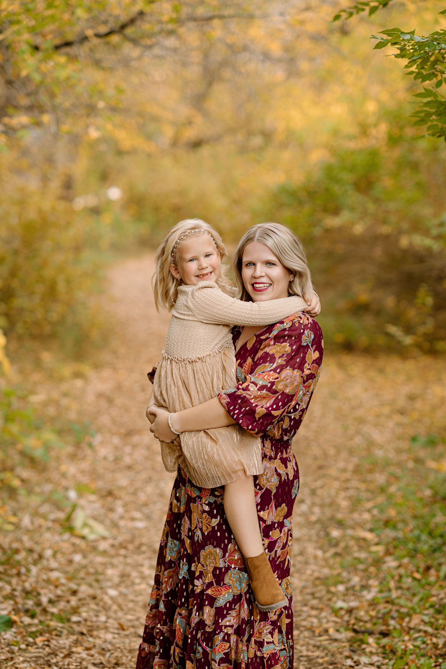 A mother holds her smiling young daughter on a leaf-covered autumn trail surrounded by golden foliage.