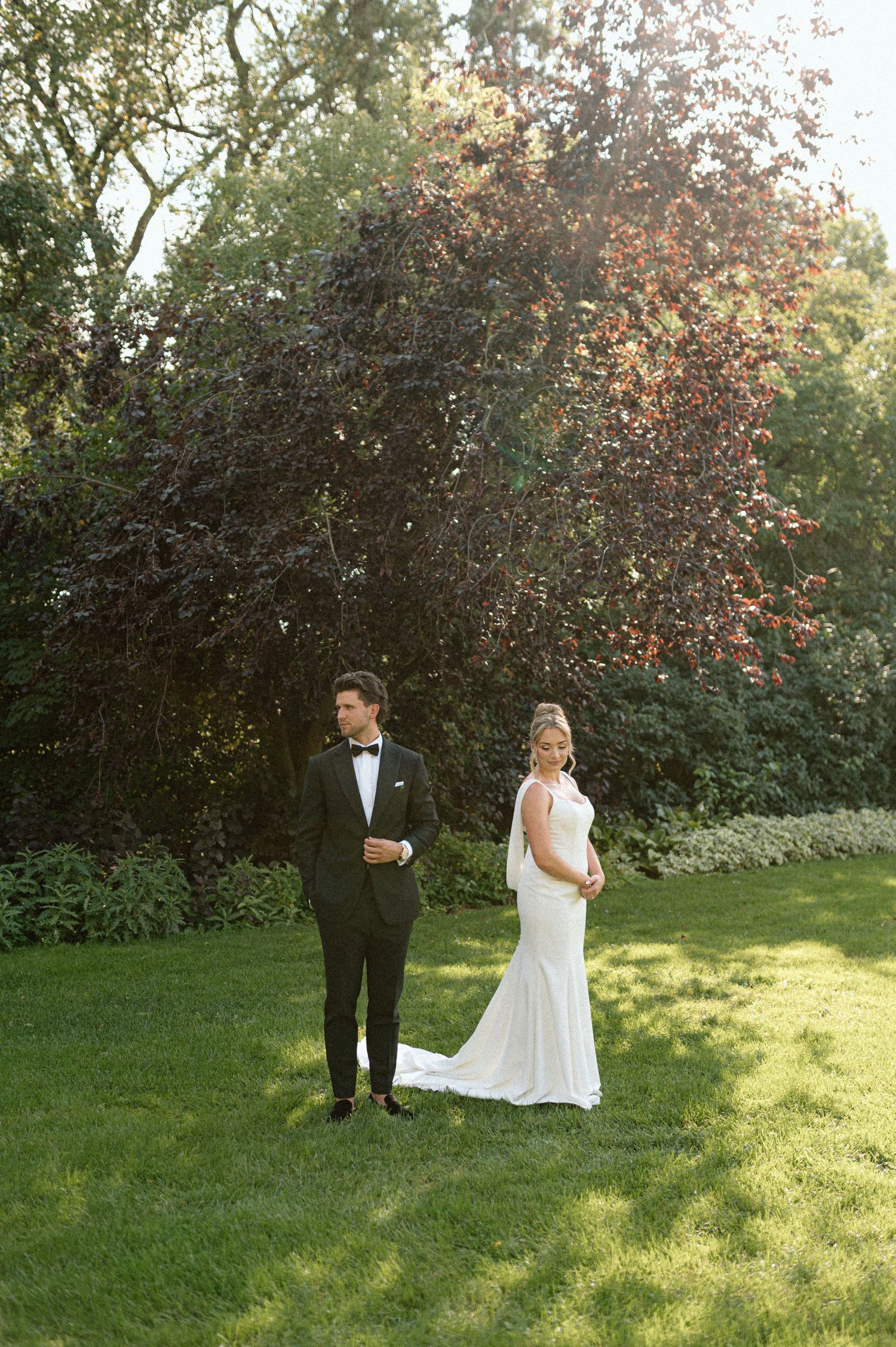 Bride in white mermaid gown and groom in tuxedo standing apart on sunlit lawn with trees.