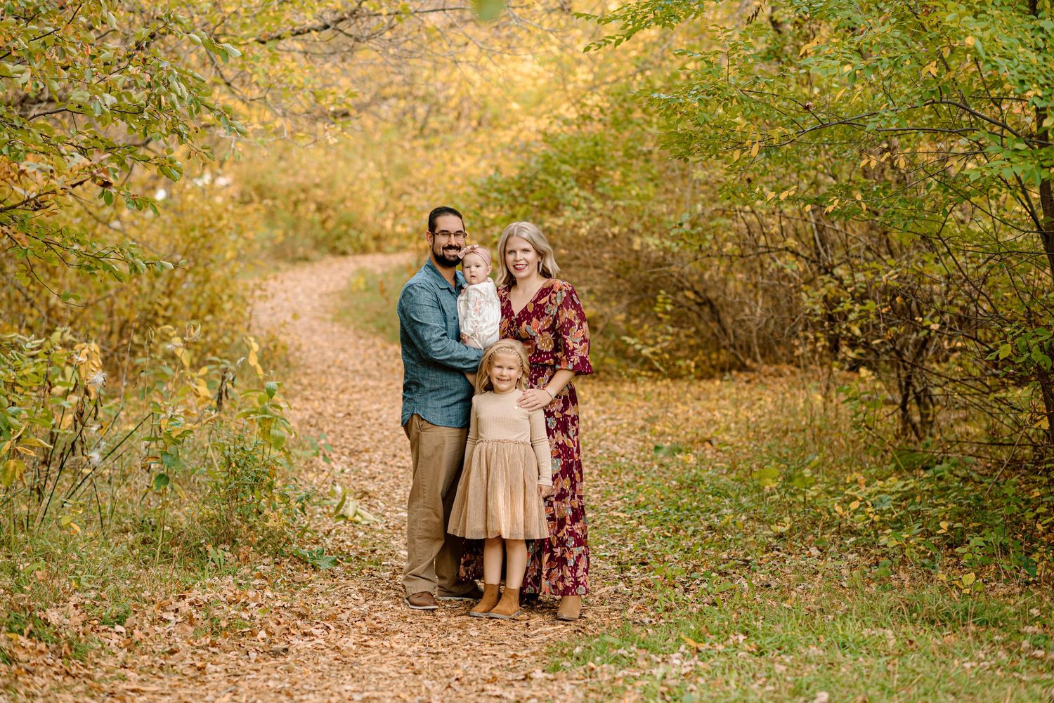 Family of three posing on a leaf-covered autumn trail surrounded by golden foliage in a scenic outdoor setting.