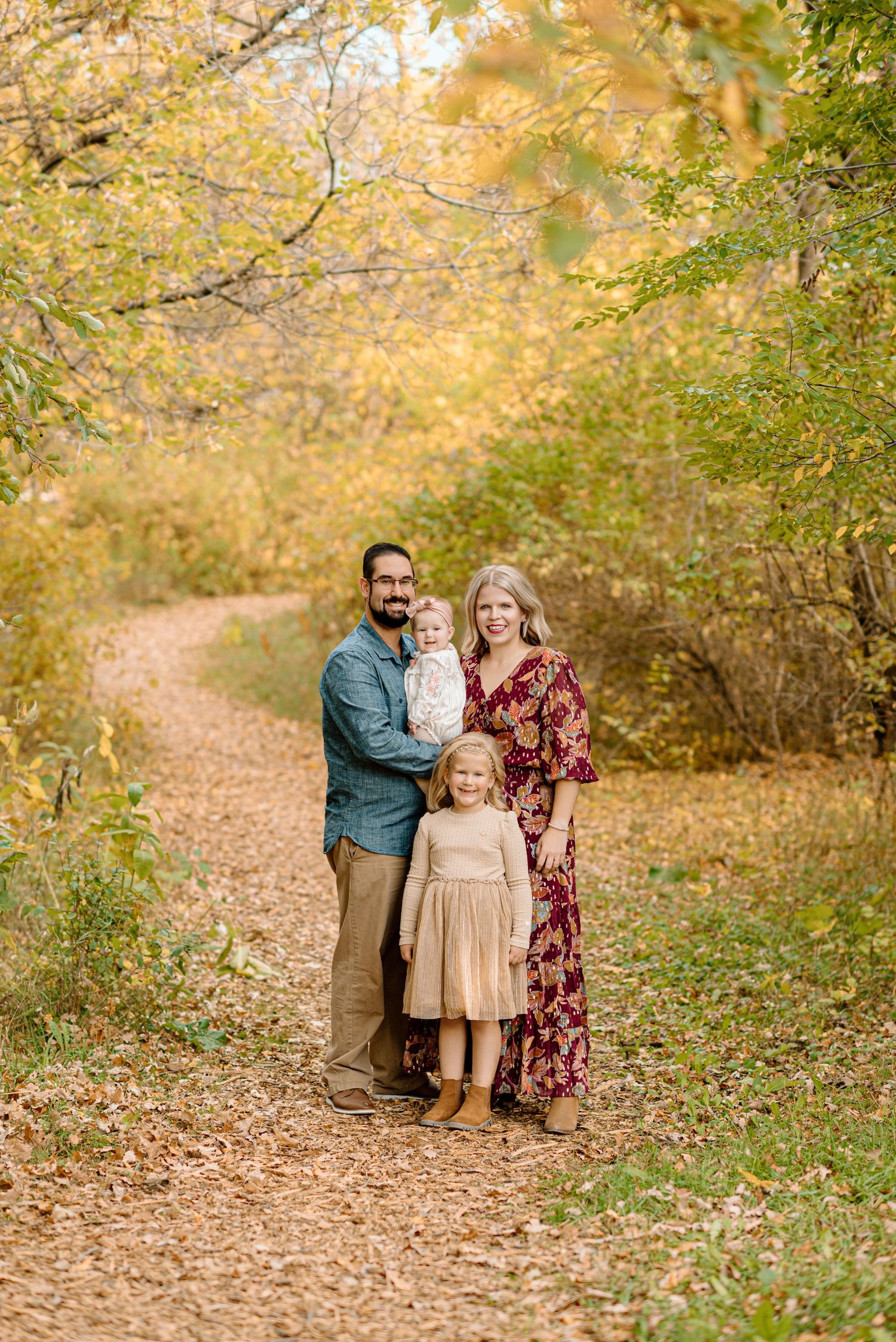 Family of four posing on a leaf-covered trail surrounded by vibrant autumn foliage in a warm fall outdoor portrait.