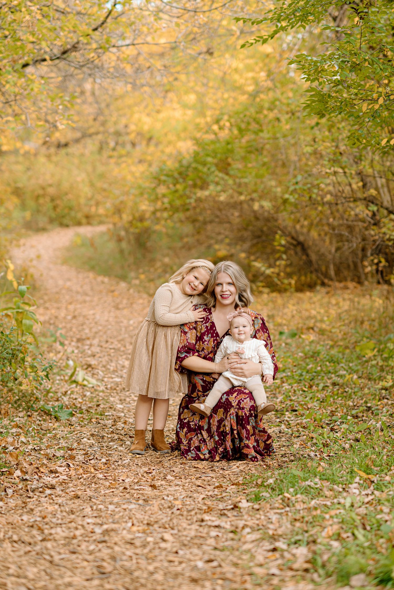 A mother sits on a fall forest path holding a baby, with a young girl leaning against her, surrounded by golden autumn foliage.