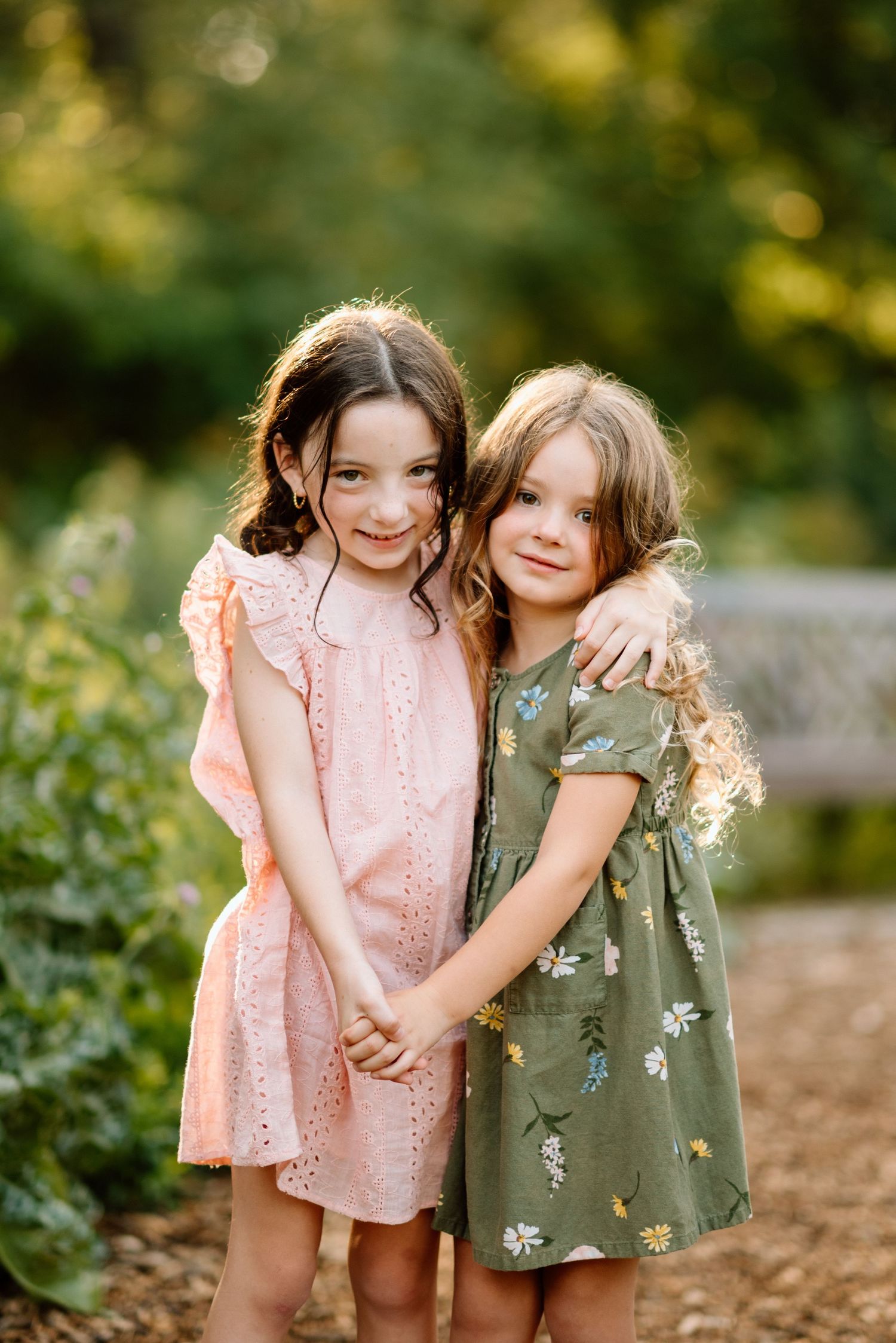 Two young sisters in pink and green floral dresses hugging each other in a sunlit garden.