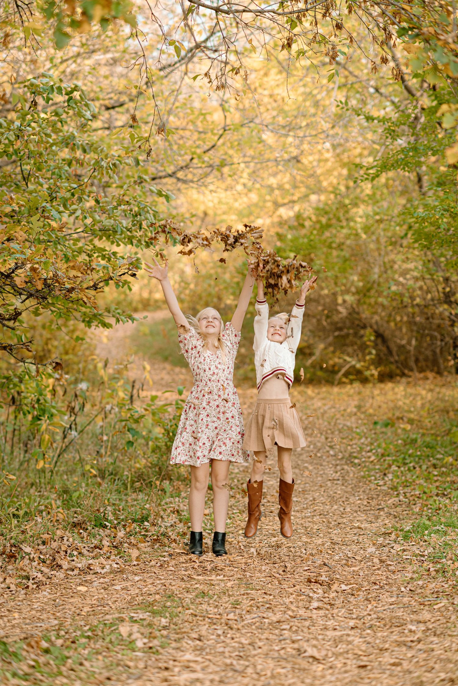 Two young girls joyfully toss colorful autumn leaves into the air on a tree-lined fall path.