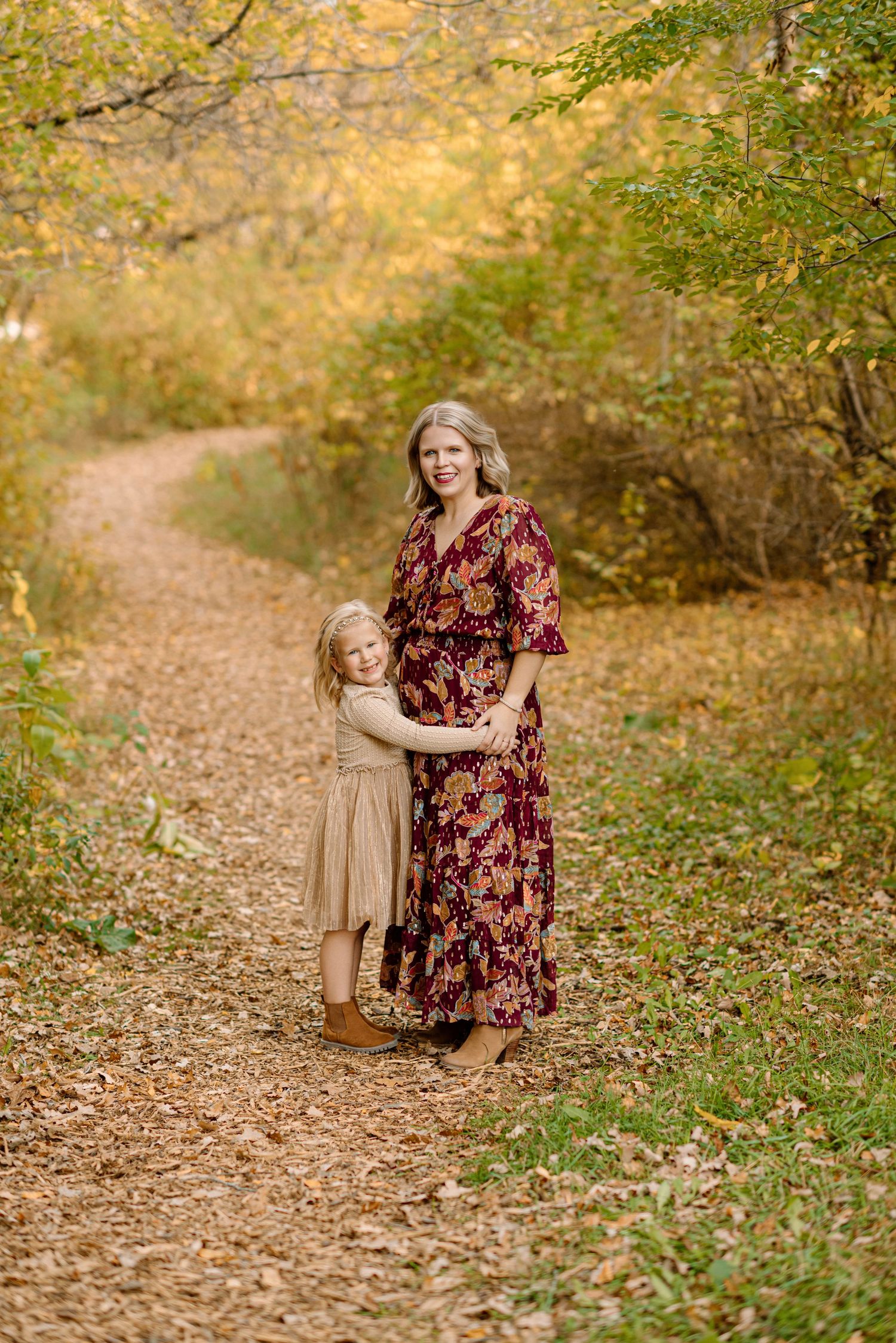 Pregnant woman in floral dress hugging young daughter on autumn leaf-covered path.