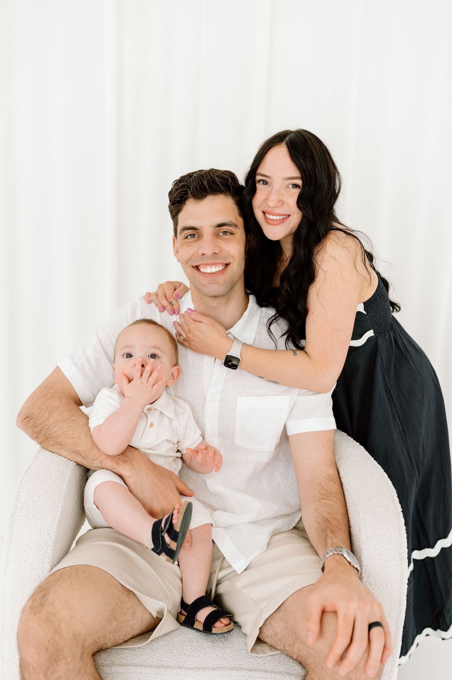 Smiling couple with their baby sitting in a white chair against a bright white backdrop.