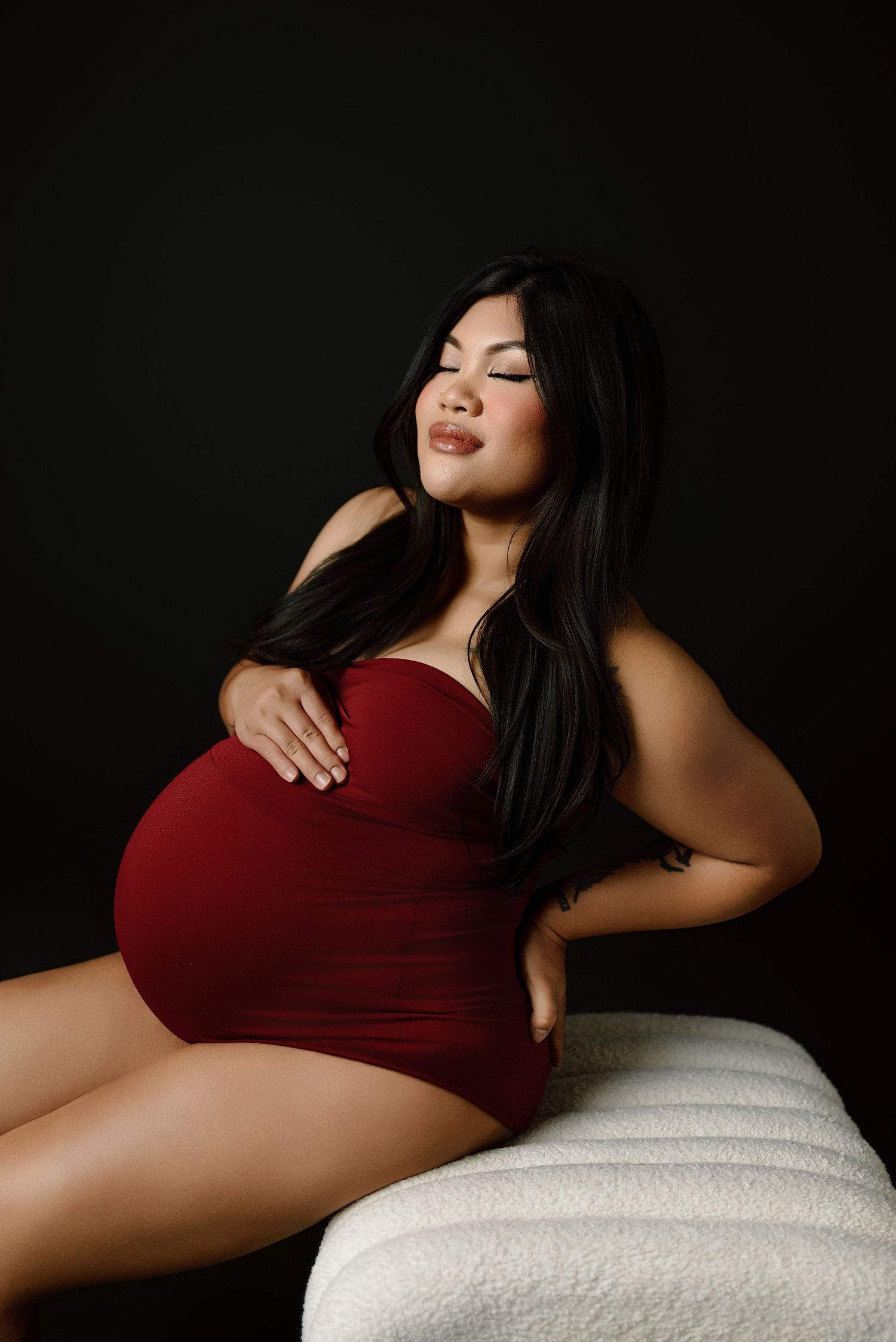 Pregnant woman in elegant red strapless dress posing gracefully against dark studio background.