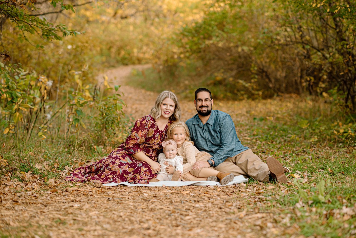 Family of four sits together on autumn leaves along a wooded trail during a fall photo session.