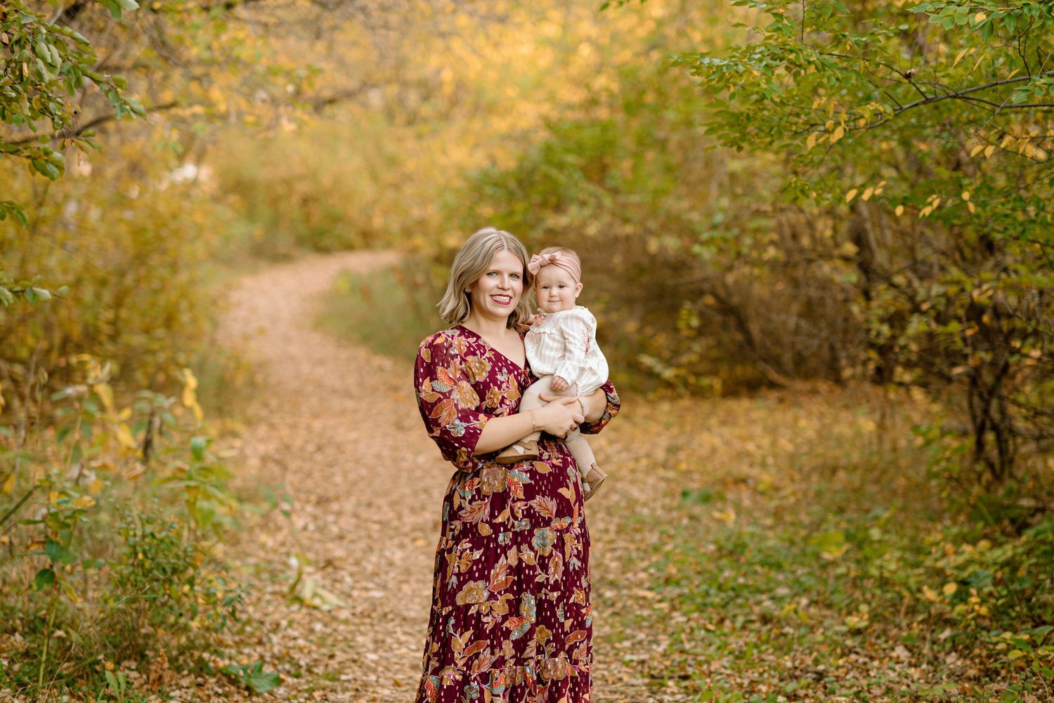 Woman in floral dress holds baby on autumn forest path surrounded by golden foliage.