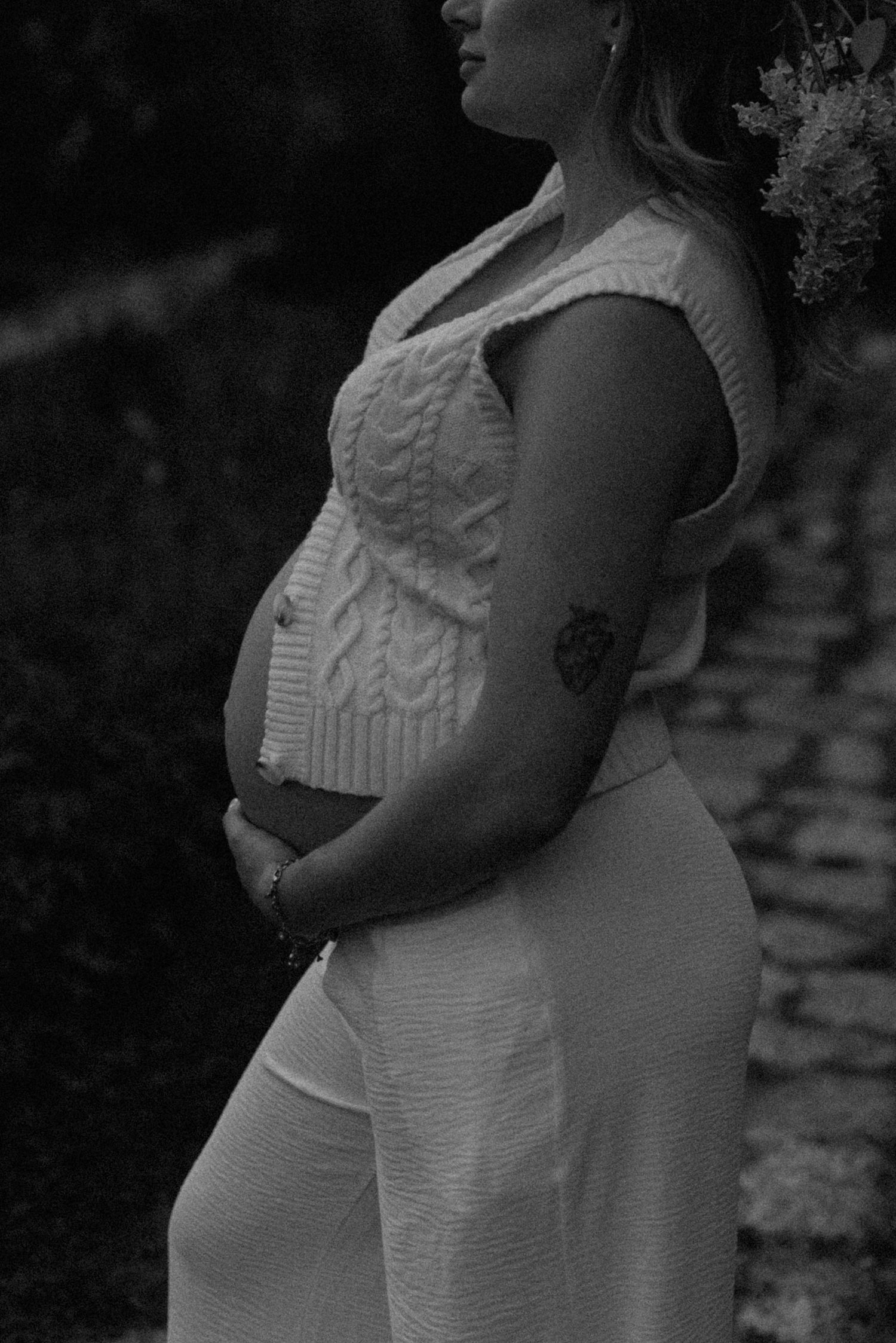 Black and white maternity photo of pregnant woman in white knit outfit cradling baby bump outdoors.