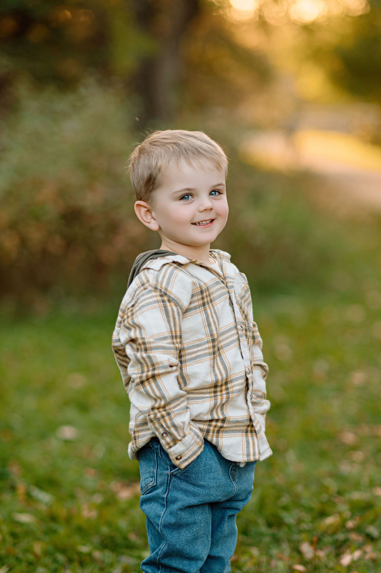 Adorable smiling toddler boy in a plaid shirt and jeans poses outdoors in golden autumn light.