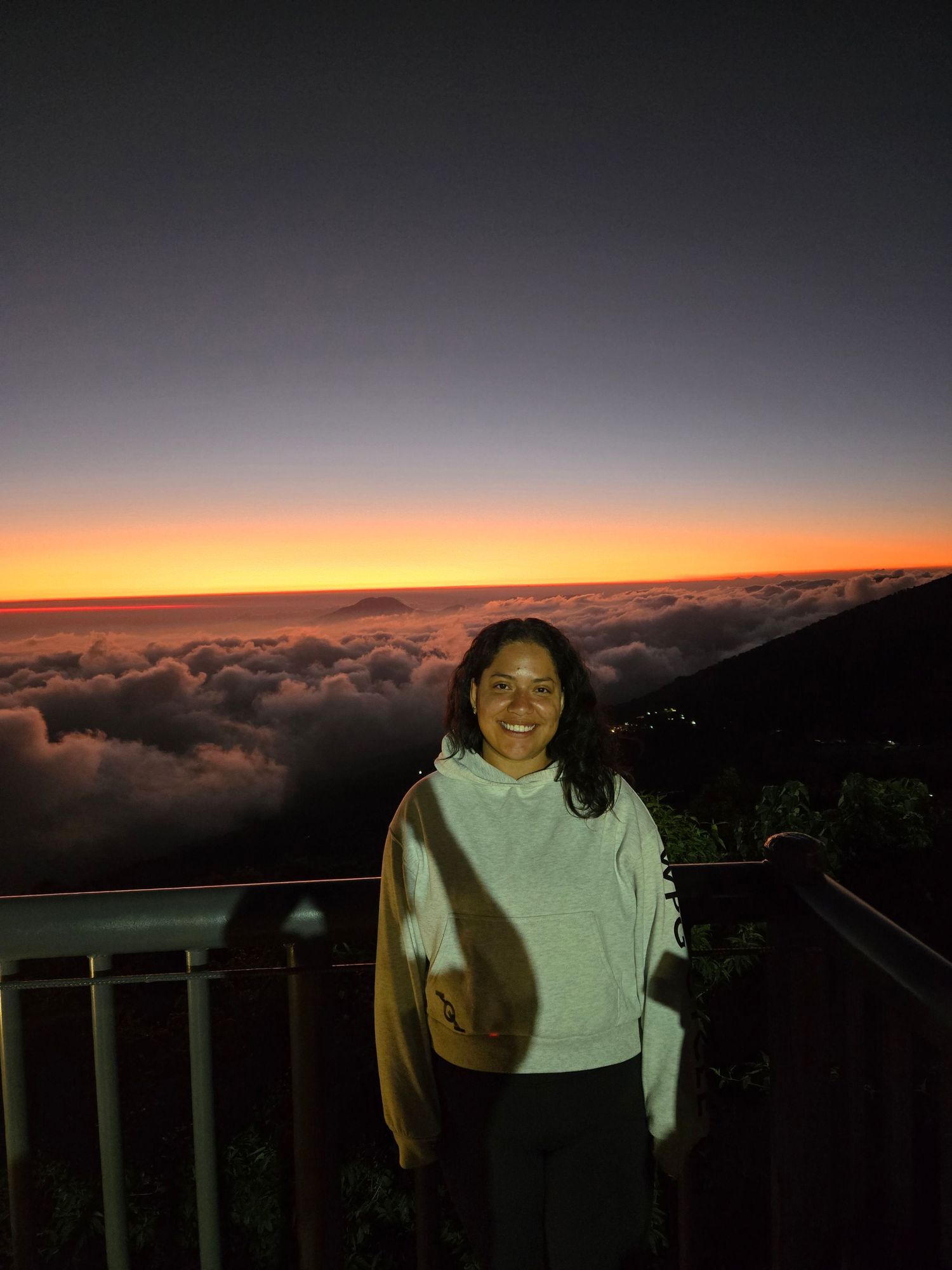 Happy woman in white hoodie standing on mountain overlook above clouds during stunning orange and red sunset.