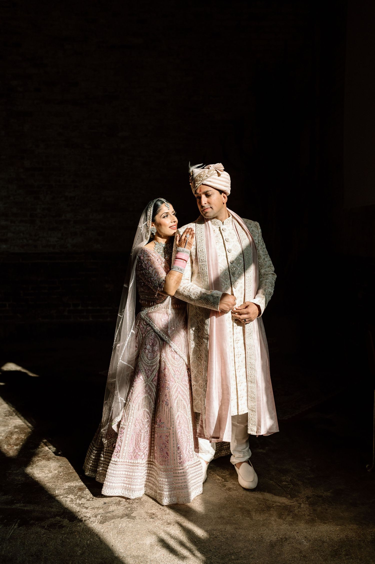 Indian wedding couple in blush lehenga and sherwani embrace in striking chiaroscuro lighting.