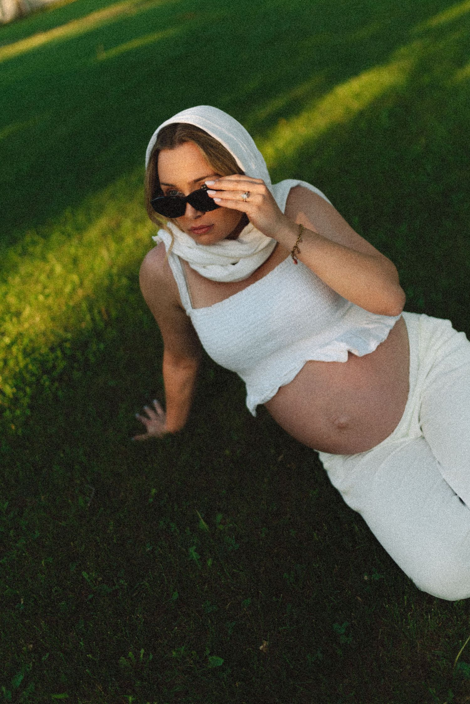 Pregnant woman in white crop top and skirt with hood and sunglasses posing on green grass in golden hour light.