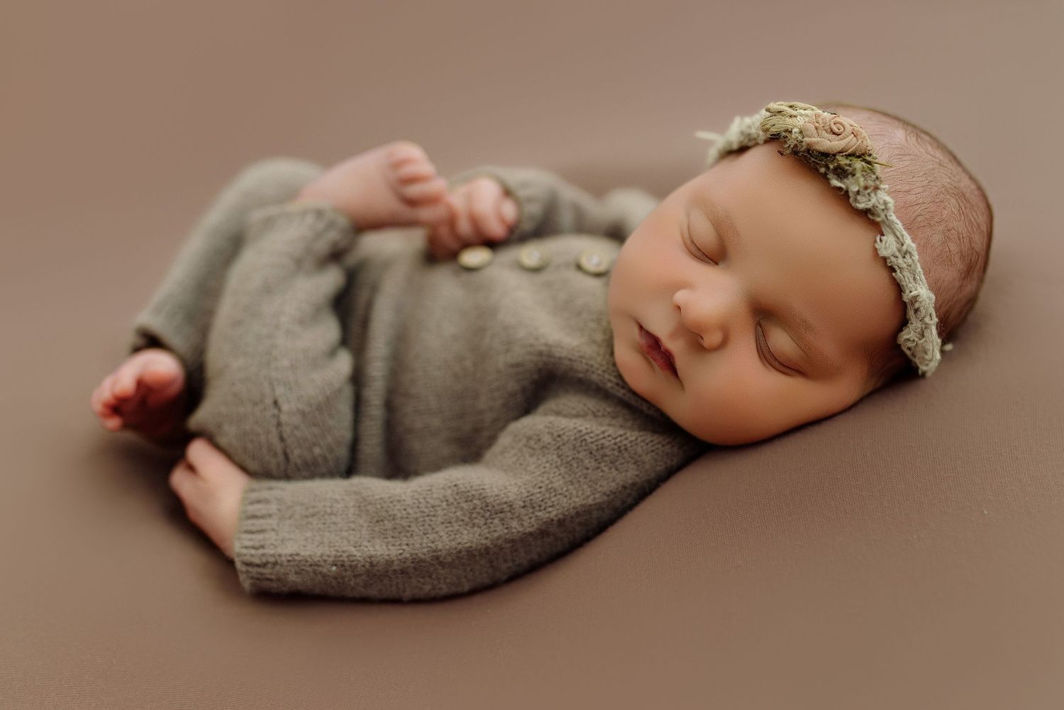 Newborn baby sleeping in knit gray romper with floral headband on brown backdrop.