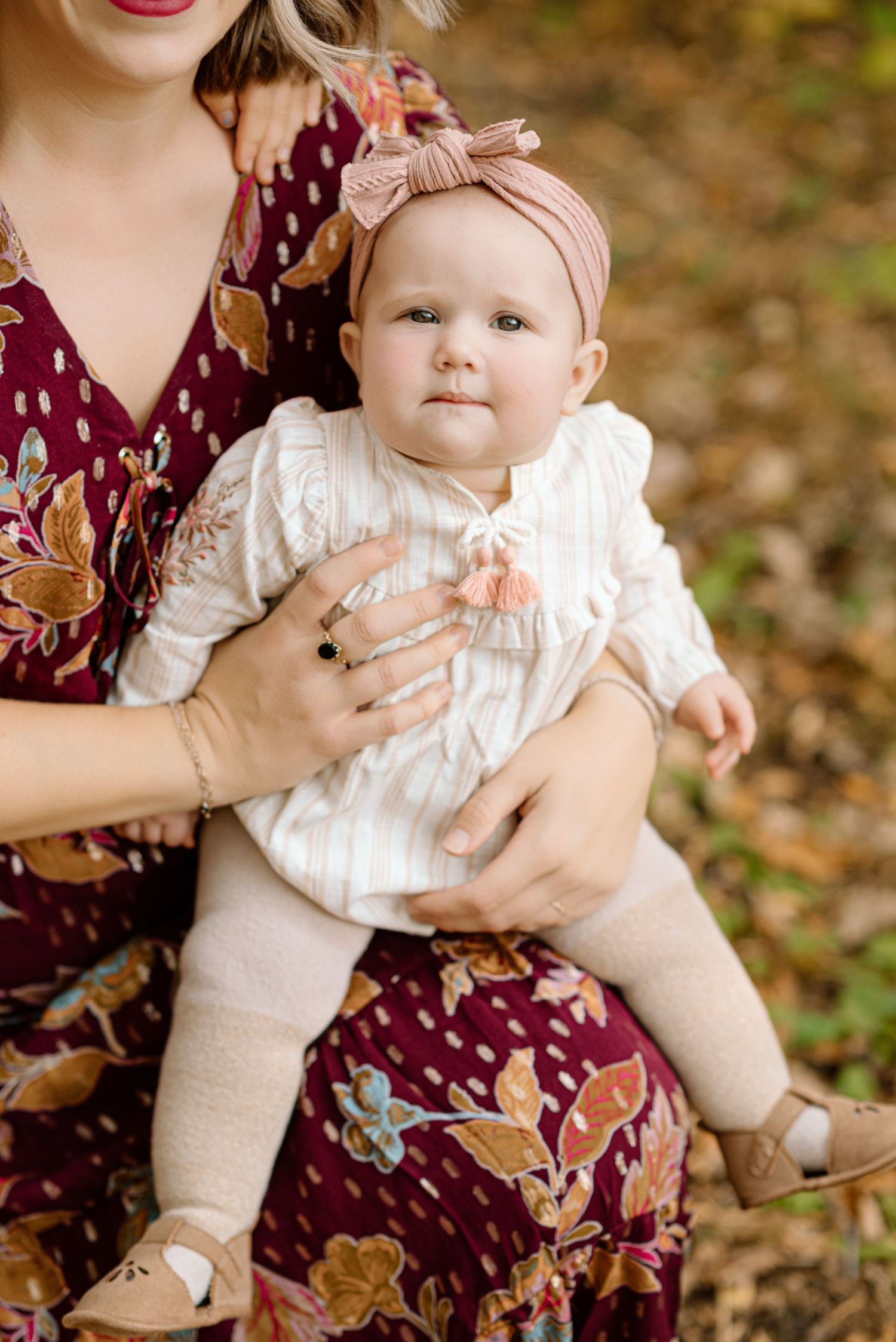 A mother in a floral dress holds her baby girl wearing a bow headband and white outfit outdoors in fall.