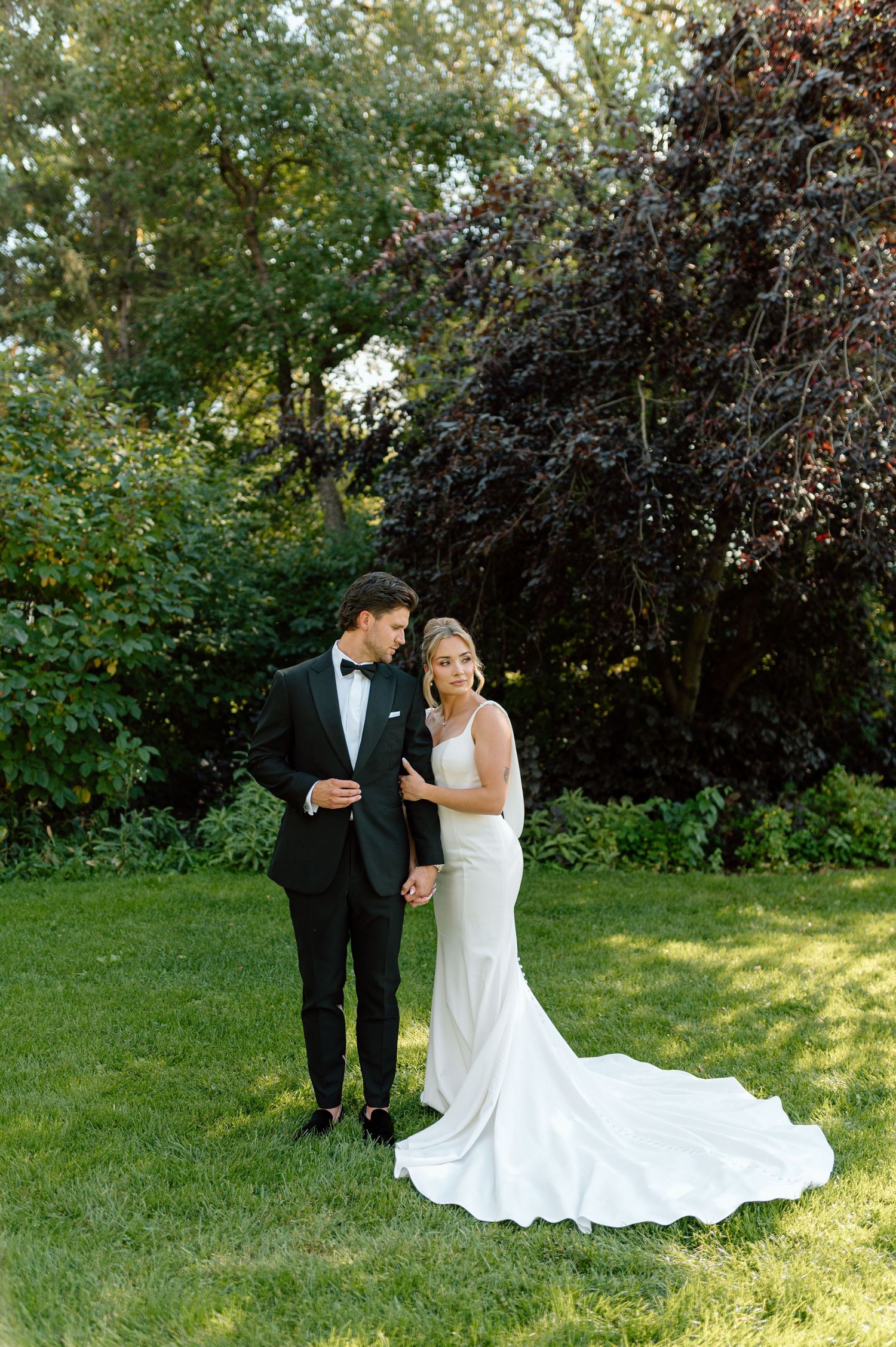 Bride in elegant white gown with train and groom in black tuxedo pose on lush green lawn.