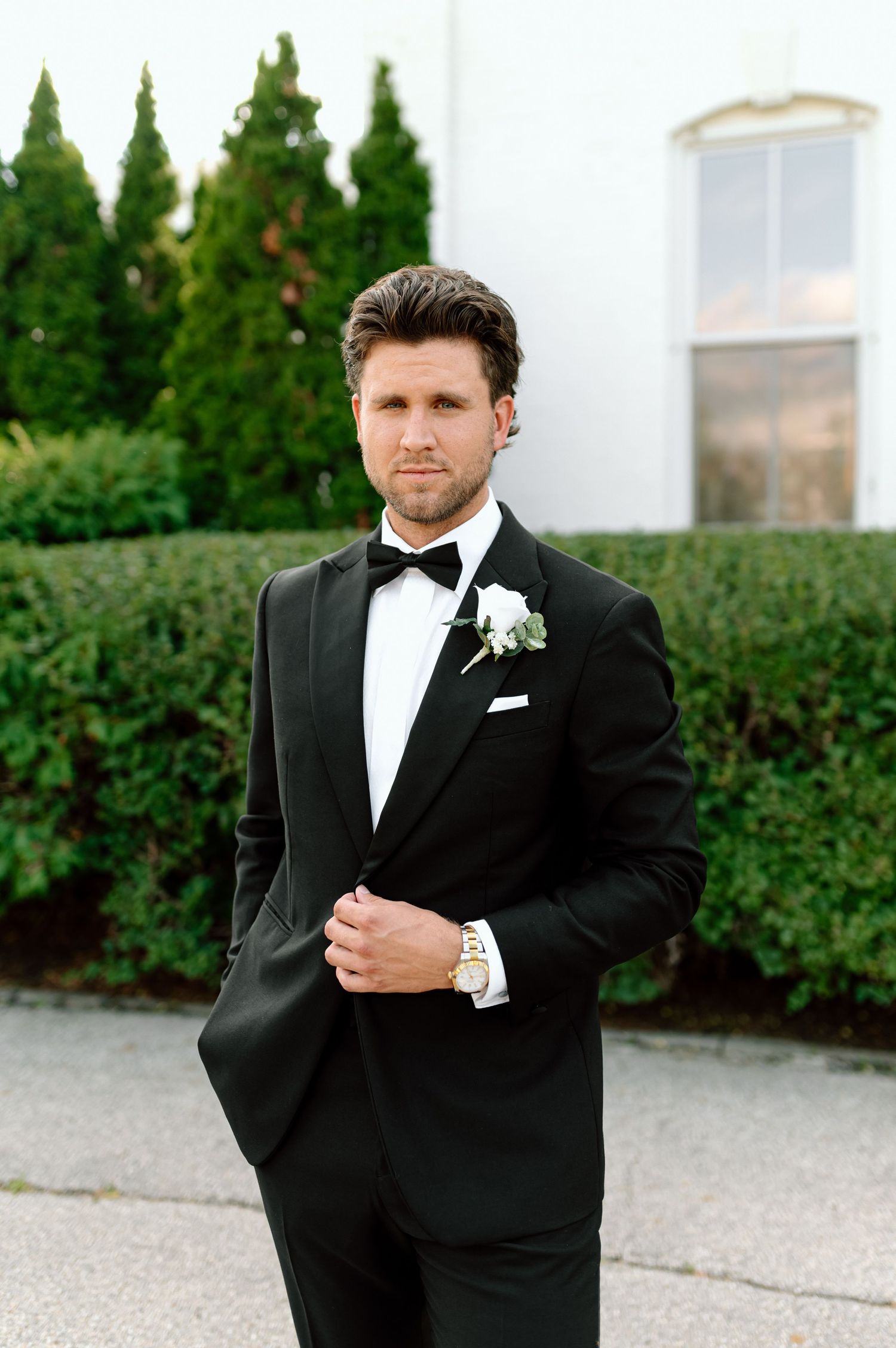 Groom in black tuxedo with bow tie and white boutonniere poses outdoors near green hedges.