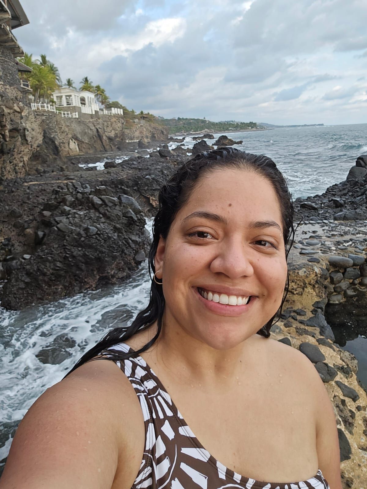 Smiling woman taking selfie on rocky coastal cliffs with ocean waves behind her.