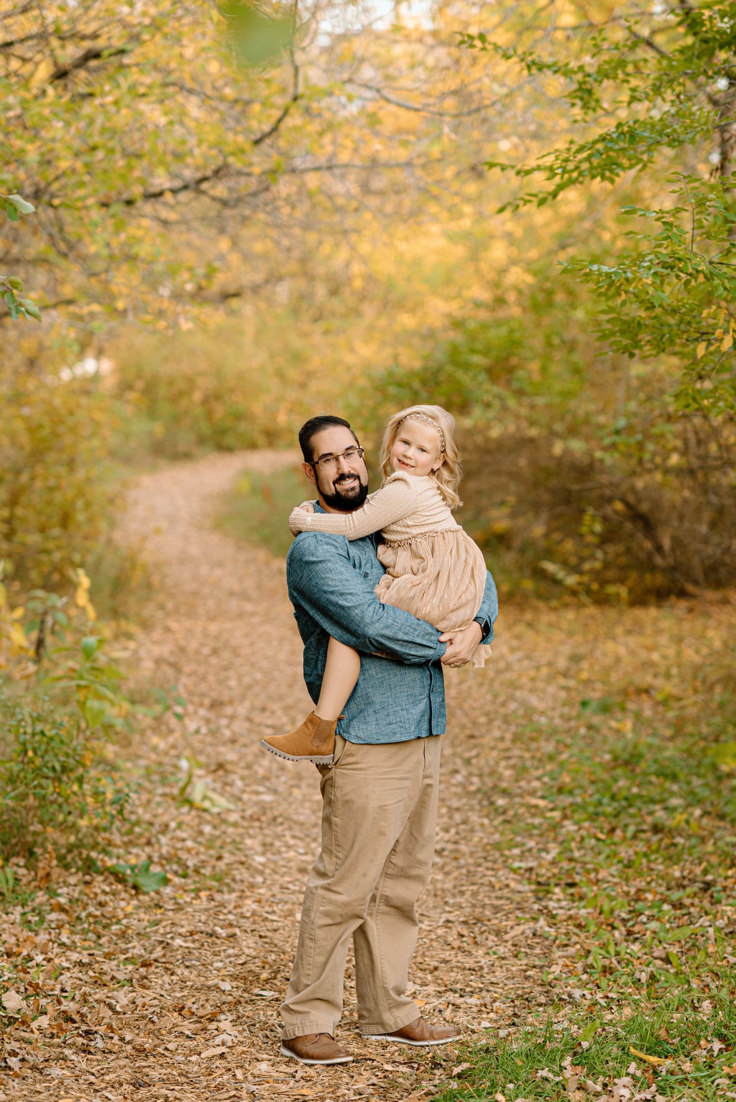 A father holds his smiling daughter on a leaf-covered autumn trail surrounded by golden foliage.
