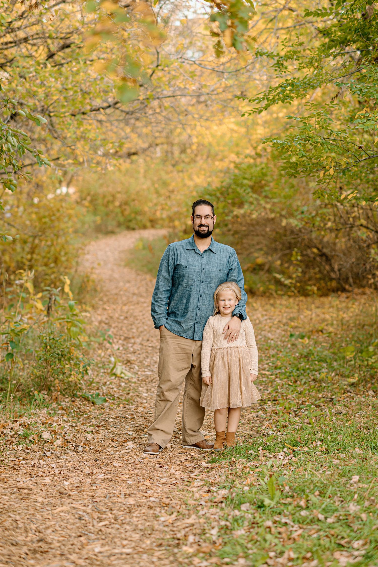 A father and young daughter pose on a leaf-covered trail surrounded by golden autumn foliage.