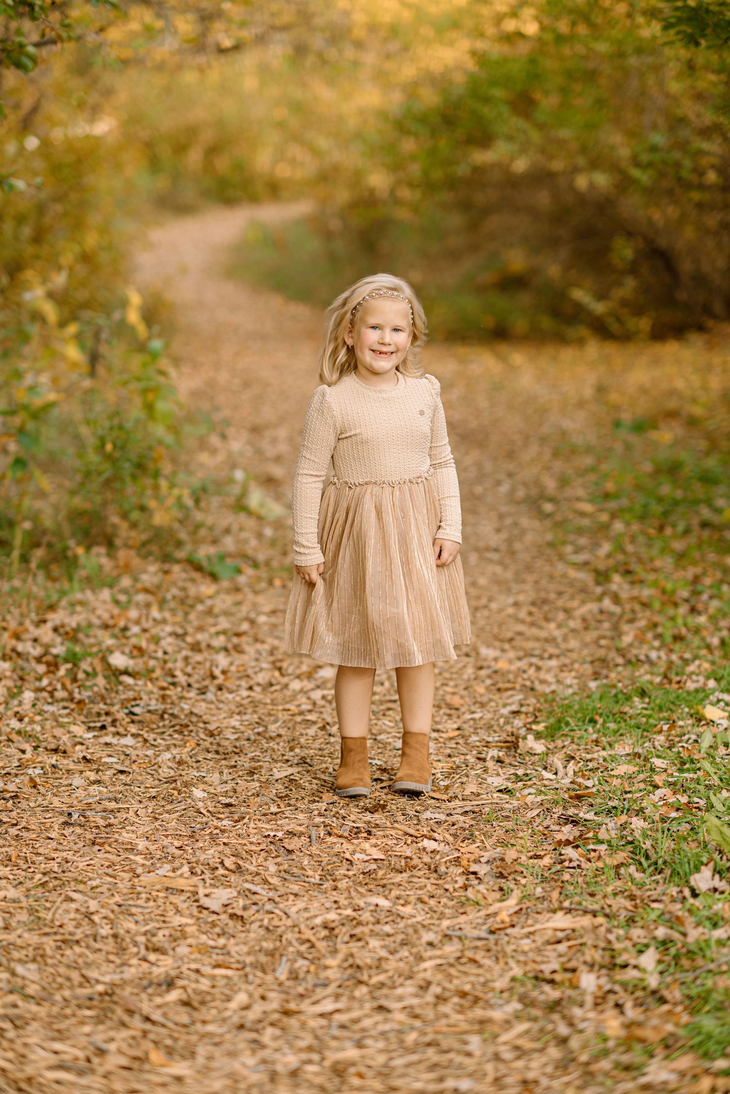 A young girl in a beige dress smiles while walking on a leaf-covered trail in autumn woods.