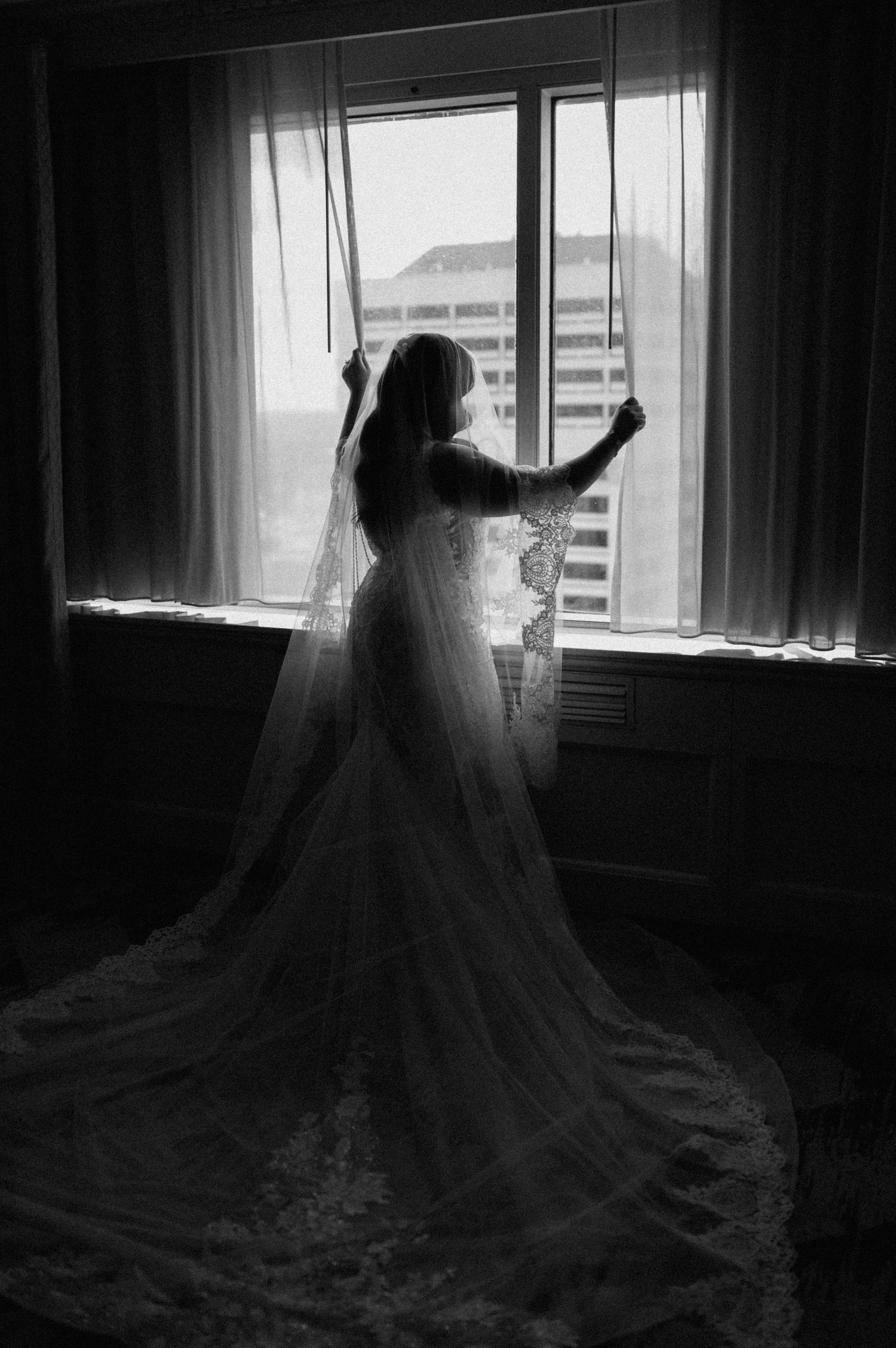 Elegant black and white silhouette of a bride in lace gown and veil standing at a hotel window.