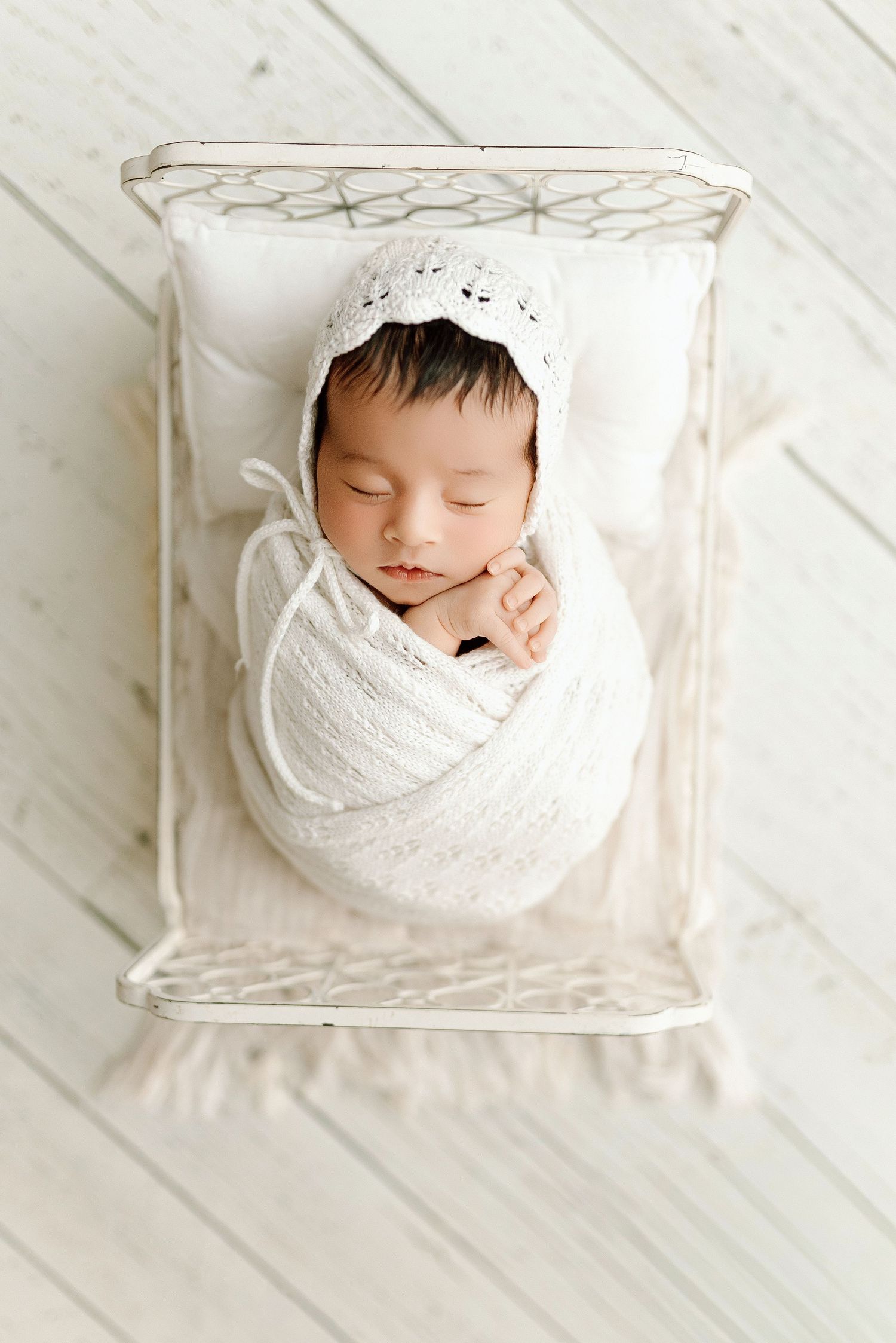 Sleeping newborn swaddled in white wrap wearing knit bonnet lying in small white metal bed.