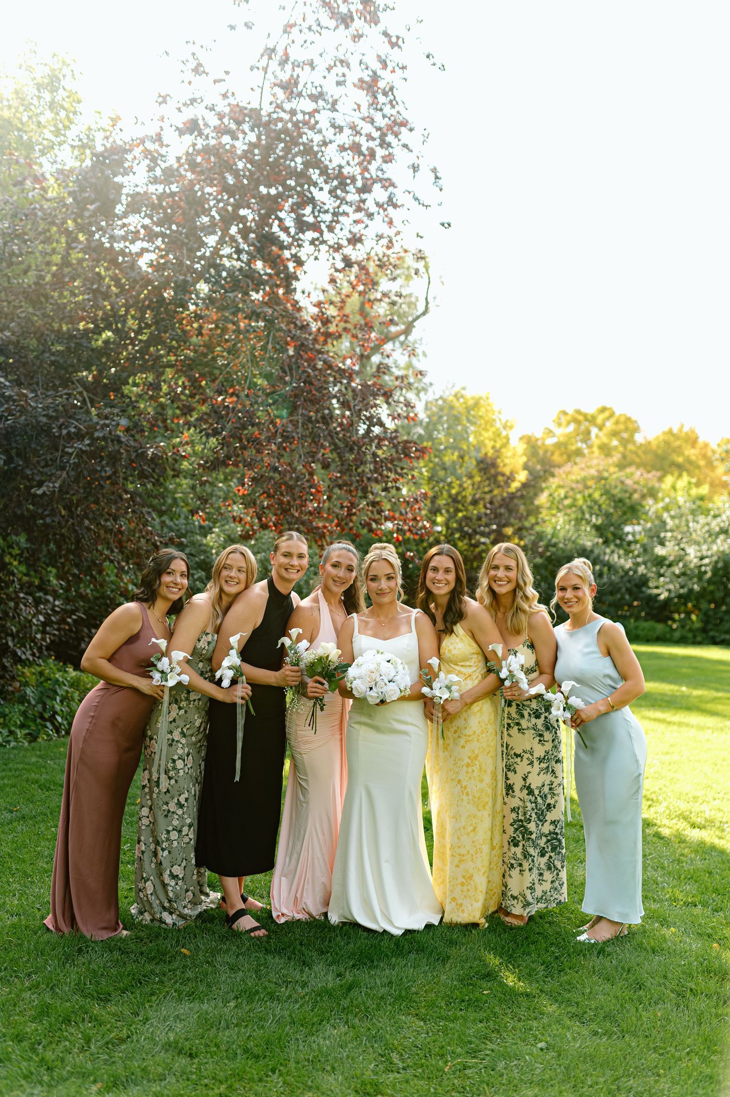 Bride poses with seven bridesmaids in mismatched colorful gowns on sunny garden lawn.