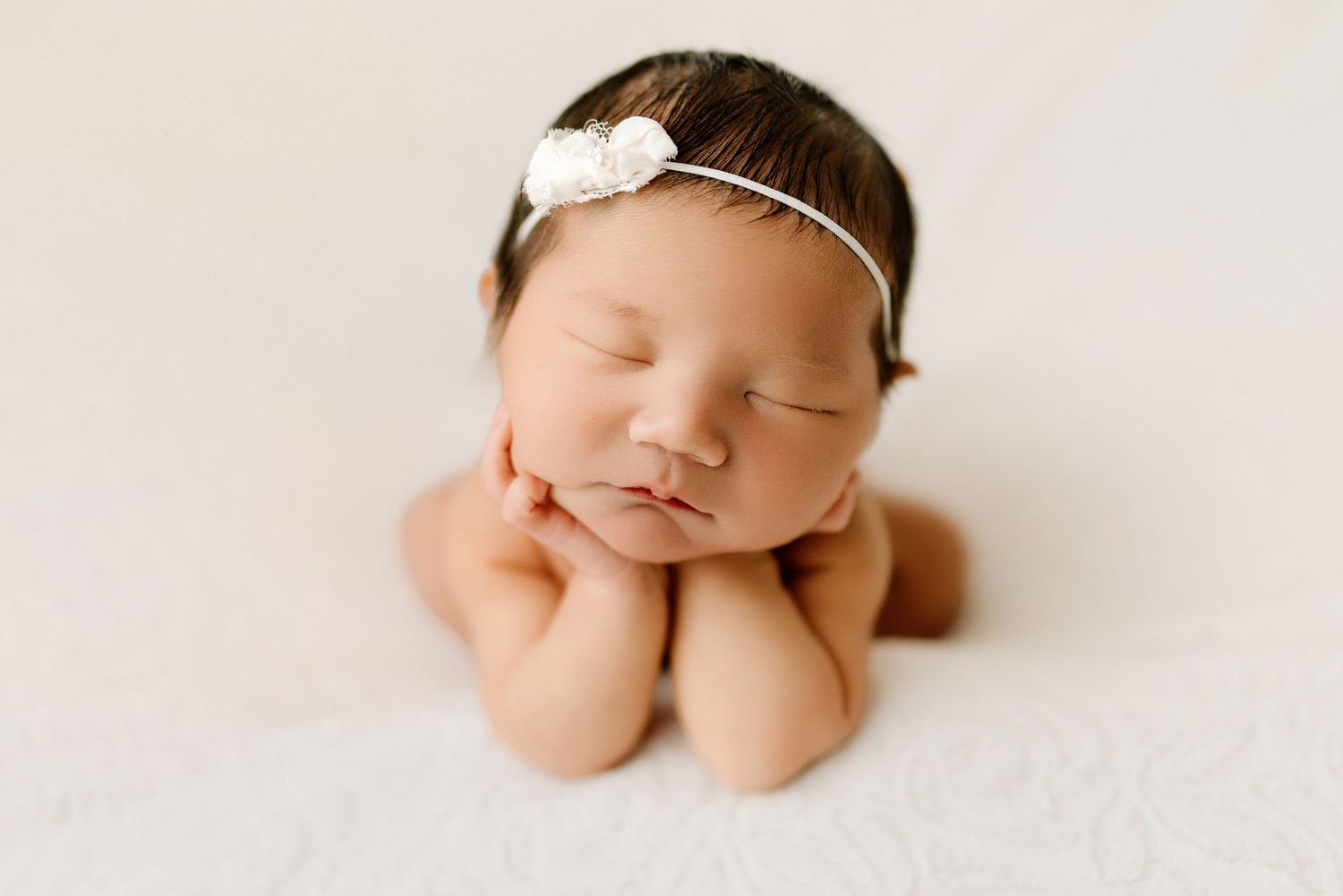 Sleeping newborn baby girl in froggy pose wearing delicate white floral headband on cream backdrop.