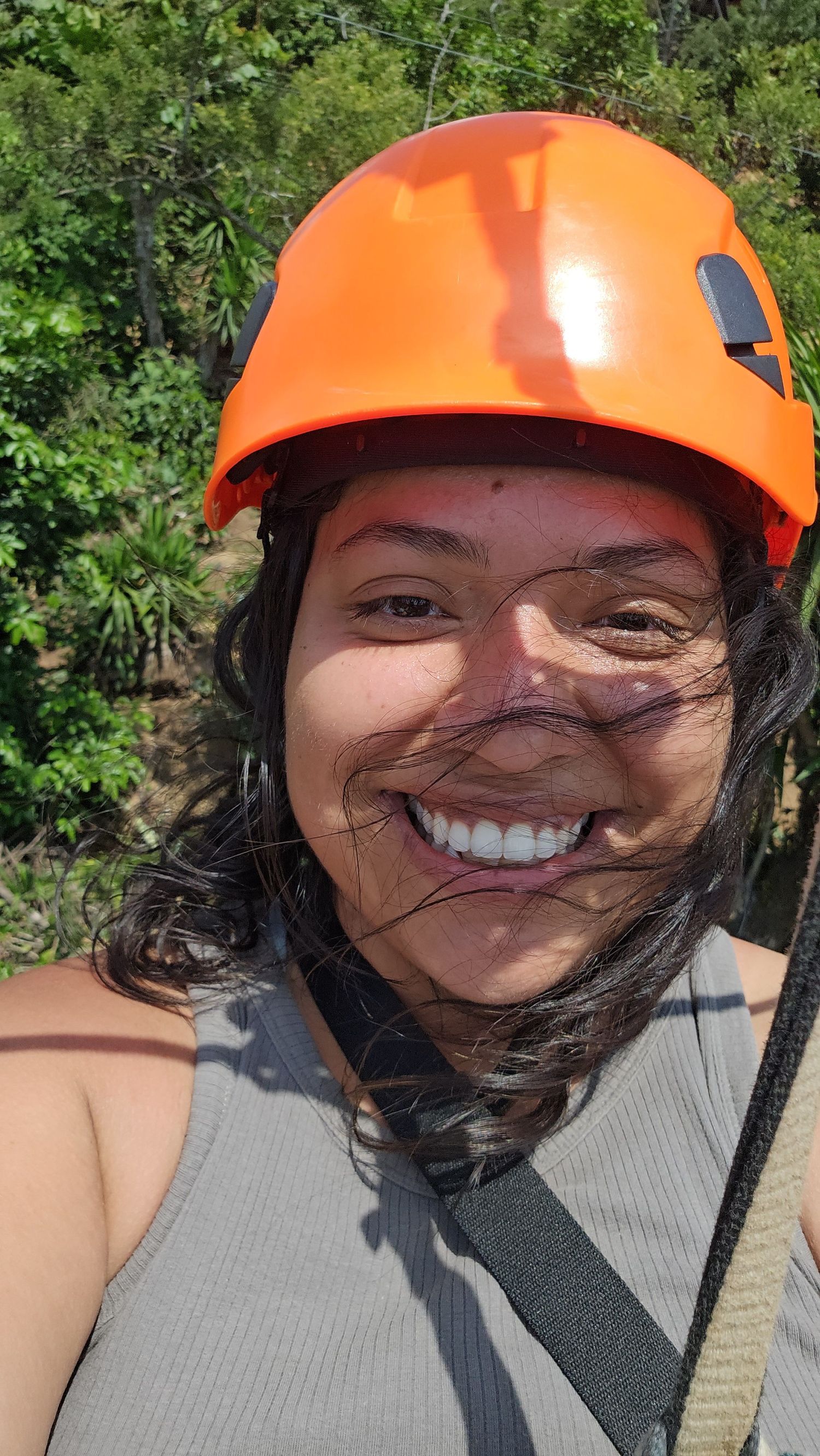 Smiling woman wearing orange safety helmet during outdoor zipline adventure activity surrounded by lush green trees.