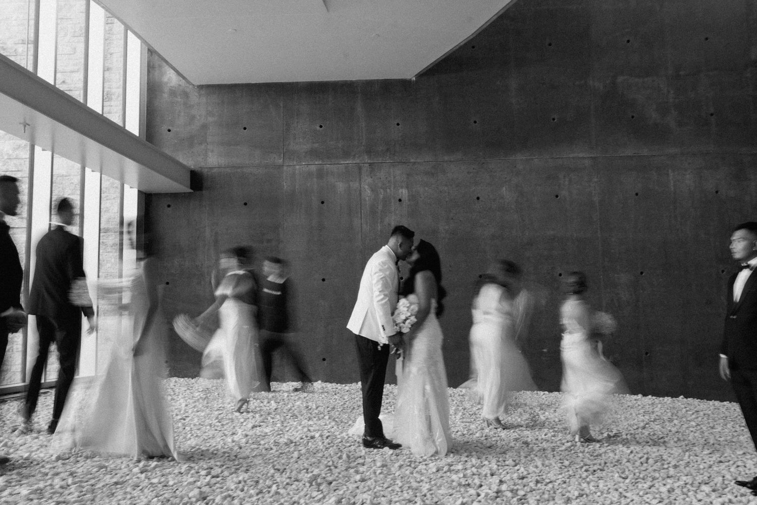 Black and white wedding photo of couple kissing surrounded by blurred guests in a modern venue.