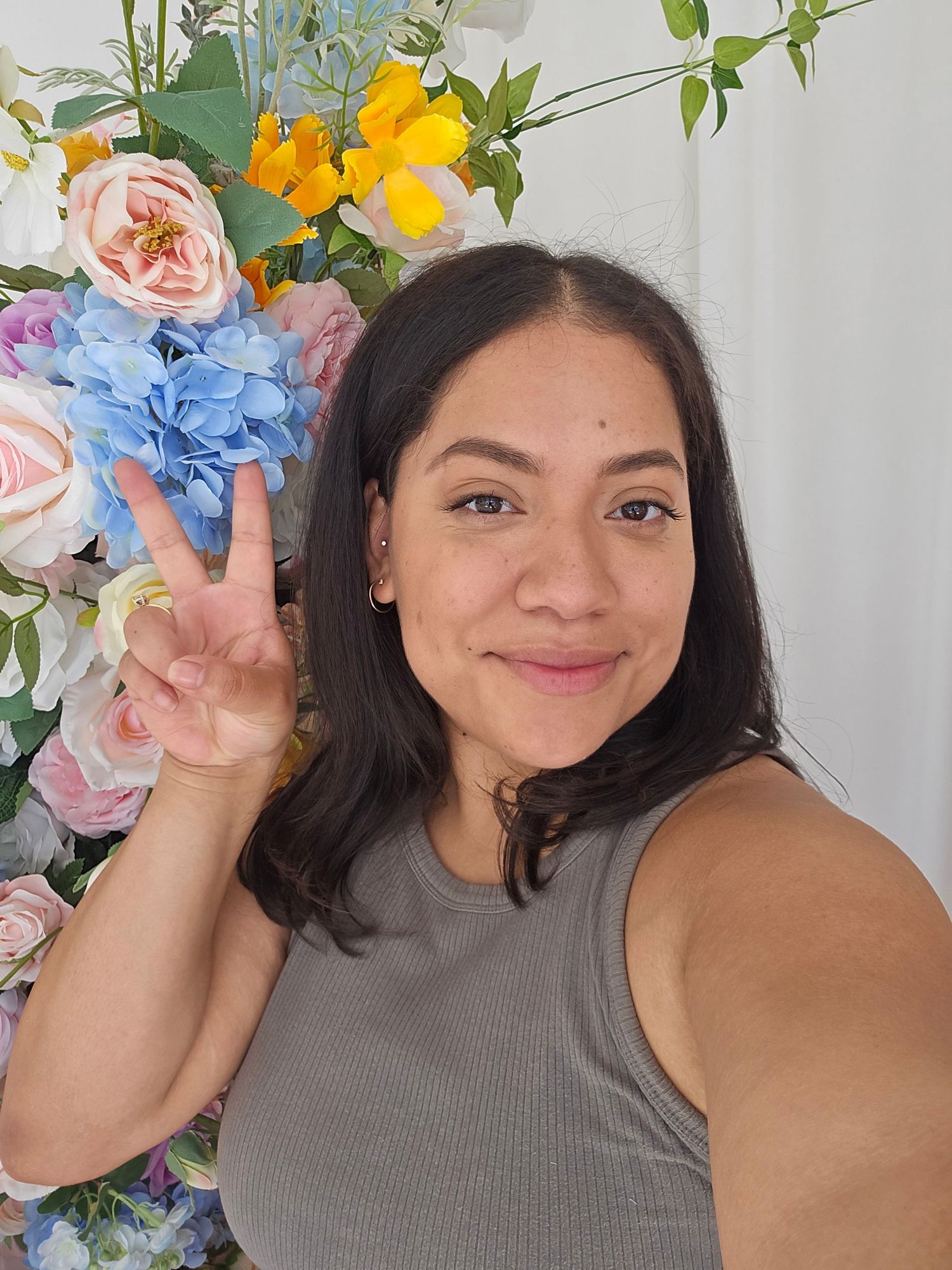Young woman taking a selfie flashing peace sign in front of colorful floral backdrop with pink blue and yellow flowers.