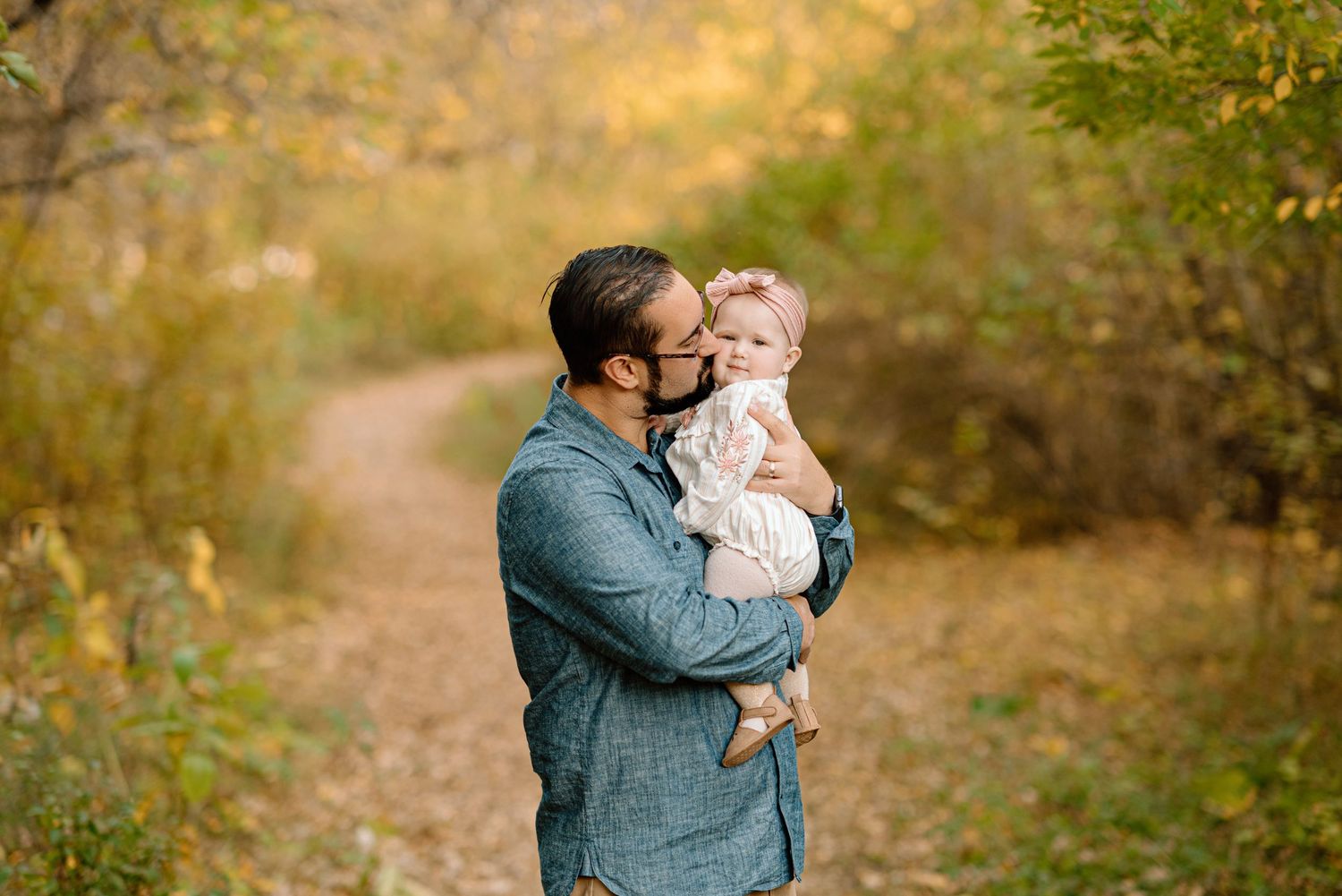 Father tenderly kissing baby daughter on a leaf-covered autumn path.