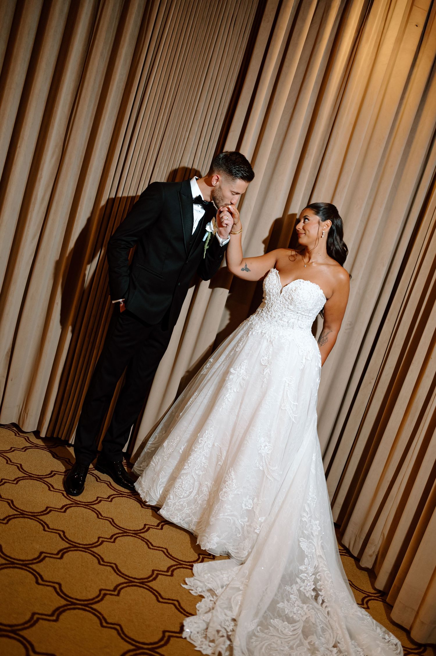Groom in black tuxedo kissing bride's hand while she smiles in a strapless lace ballgown at their wedding.