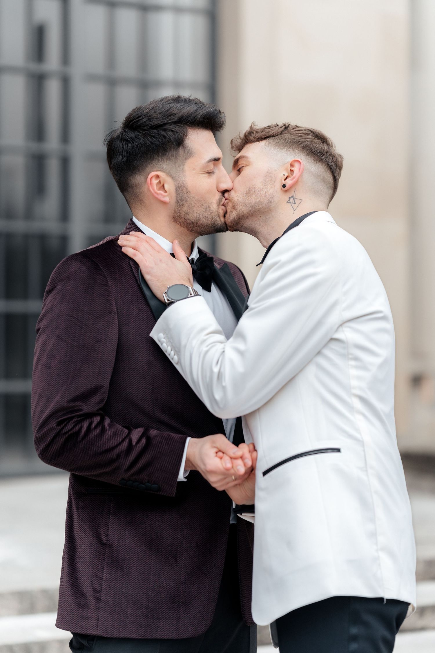 A romantic kiss between two people wearing formal attire, one in a burgundy jacket and one in white, standing outdoors.
