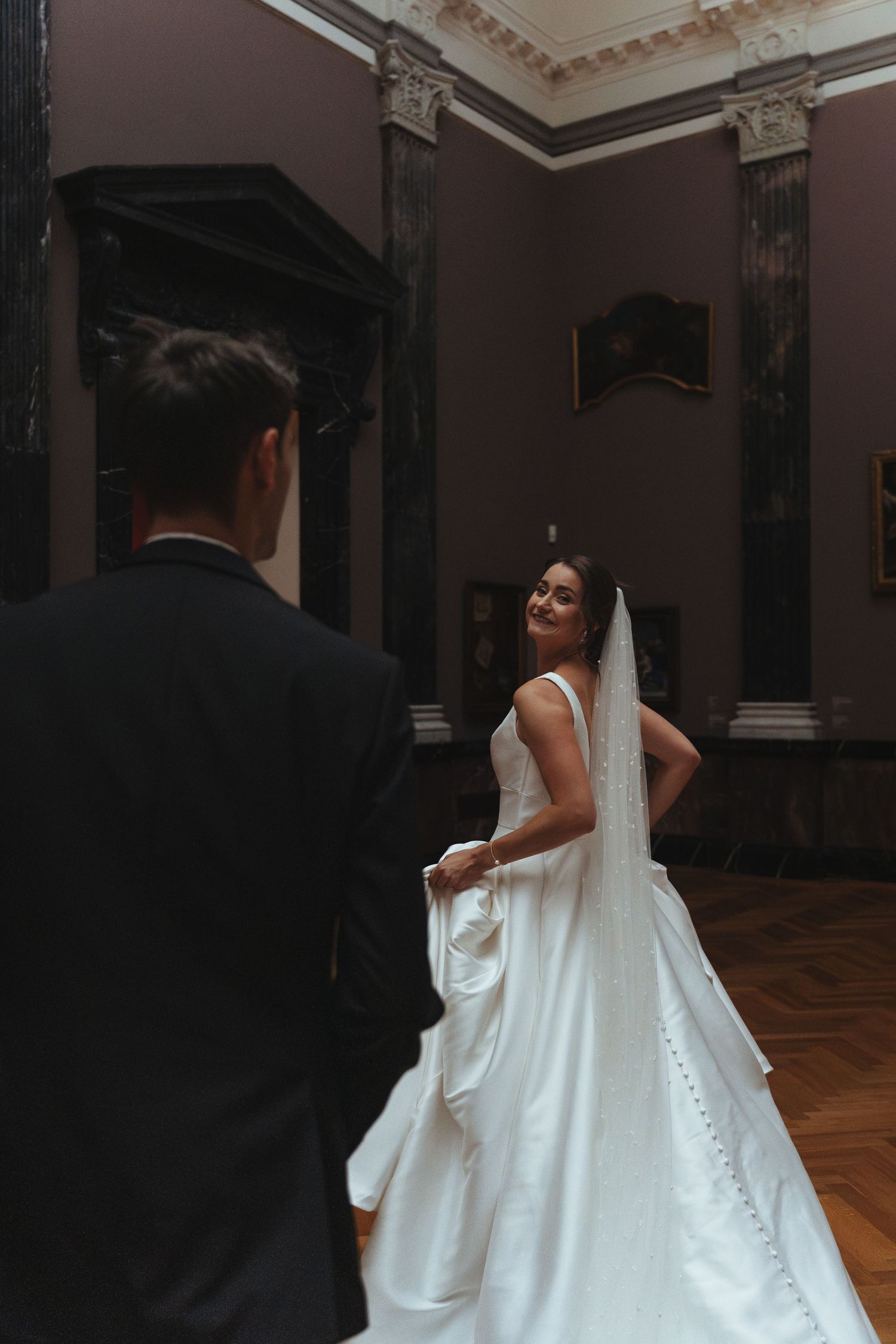 A bride in a sleek white gown poses playfully in an ornate museum gallery with dark walls and columns.