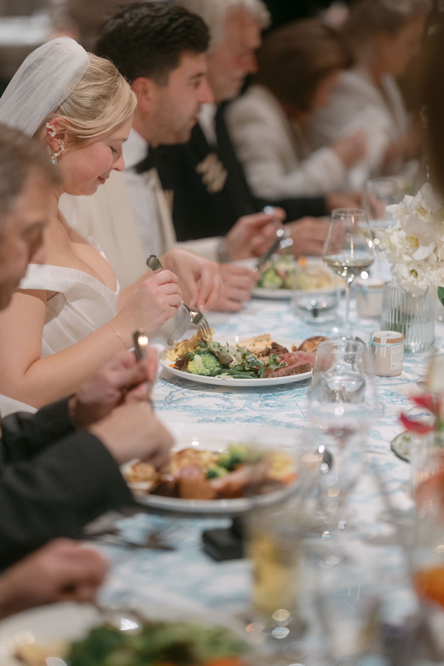 Wedding guests enjoy an elegant dinner reception with plated meals and wine glasses at a formally set table.