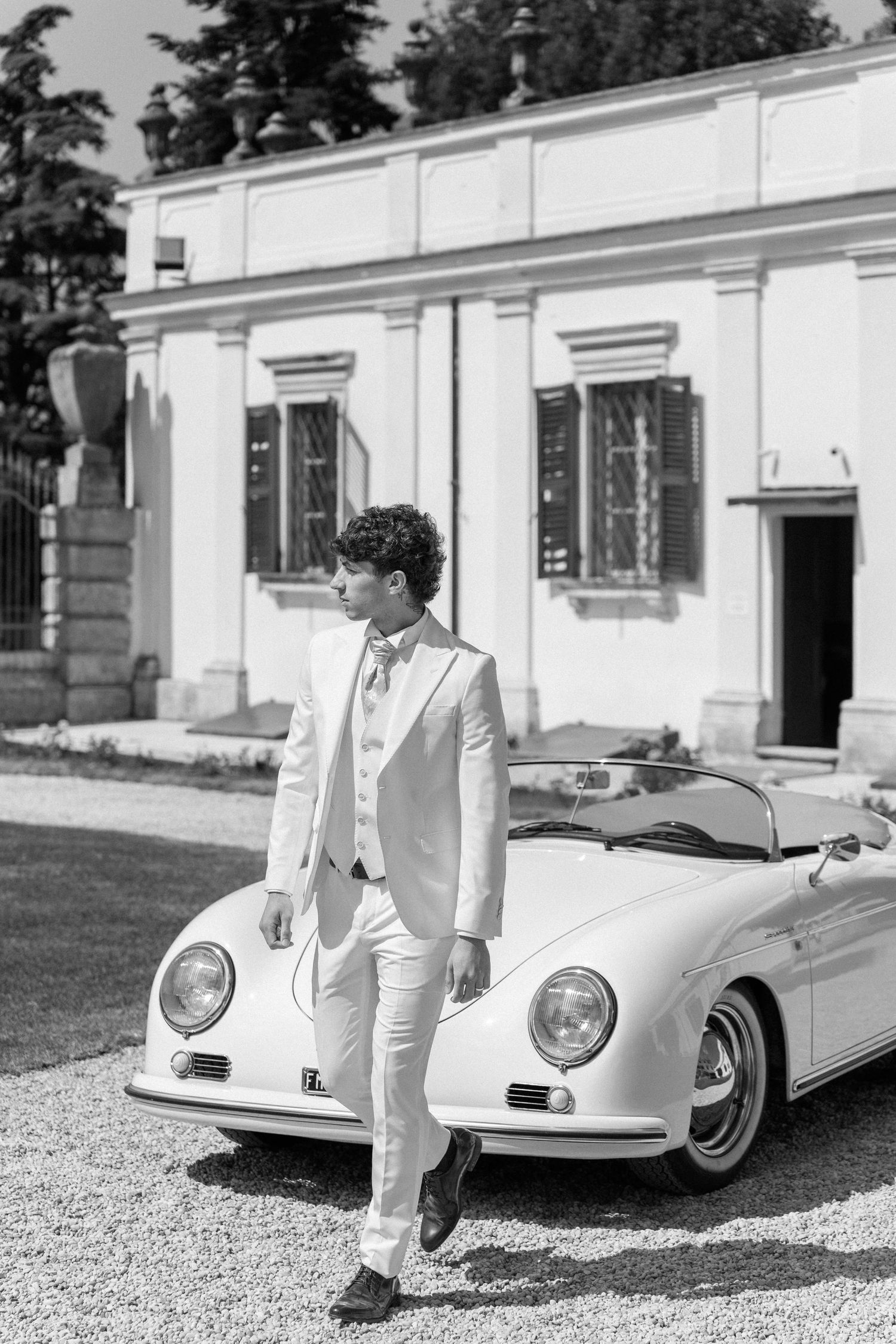 Black and white photo of a vintage Porsche 356 Speedster convertible parked in front of an elegant white building. Villa Mosconi Bertani