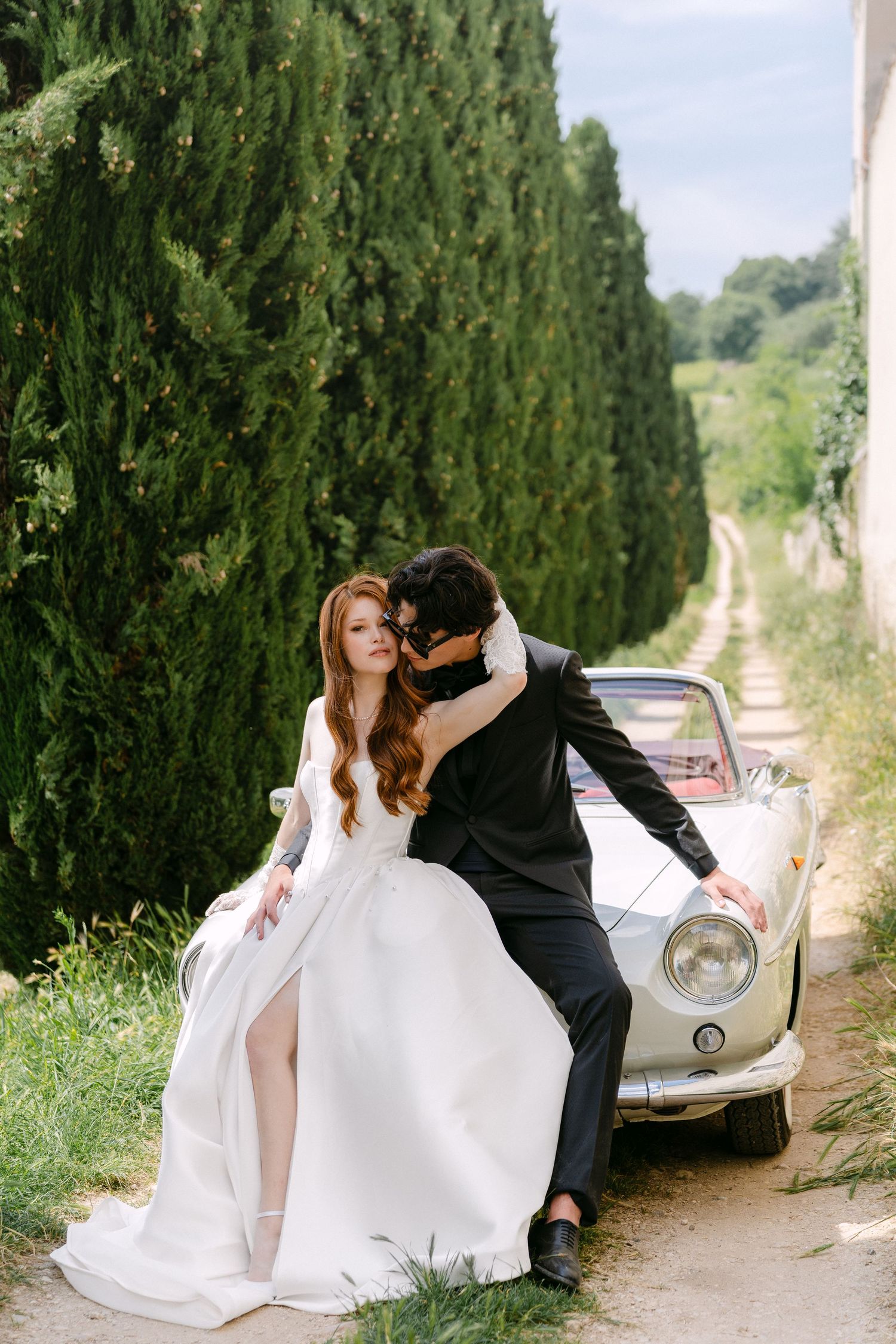 A romantic wedding couple poses along a cypress tree-lined path in Italy with a vintage car in the background.