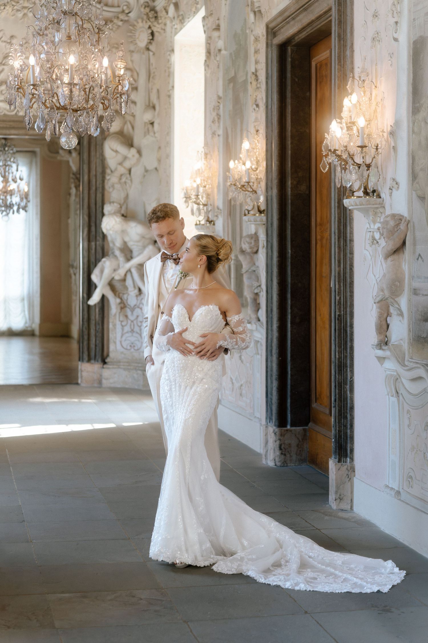 Elegant wedding couple poses in ornate baroque palace hallway with crystal chandeliers and decorative white walls. Schloss Ludwigsburg