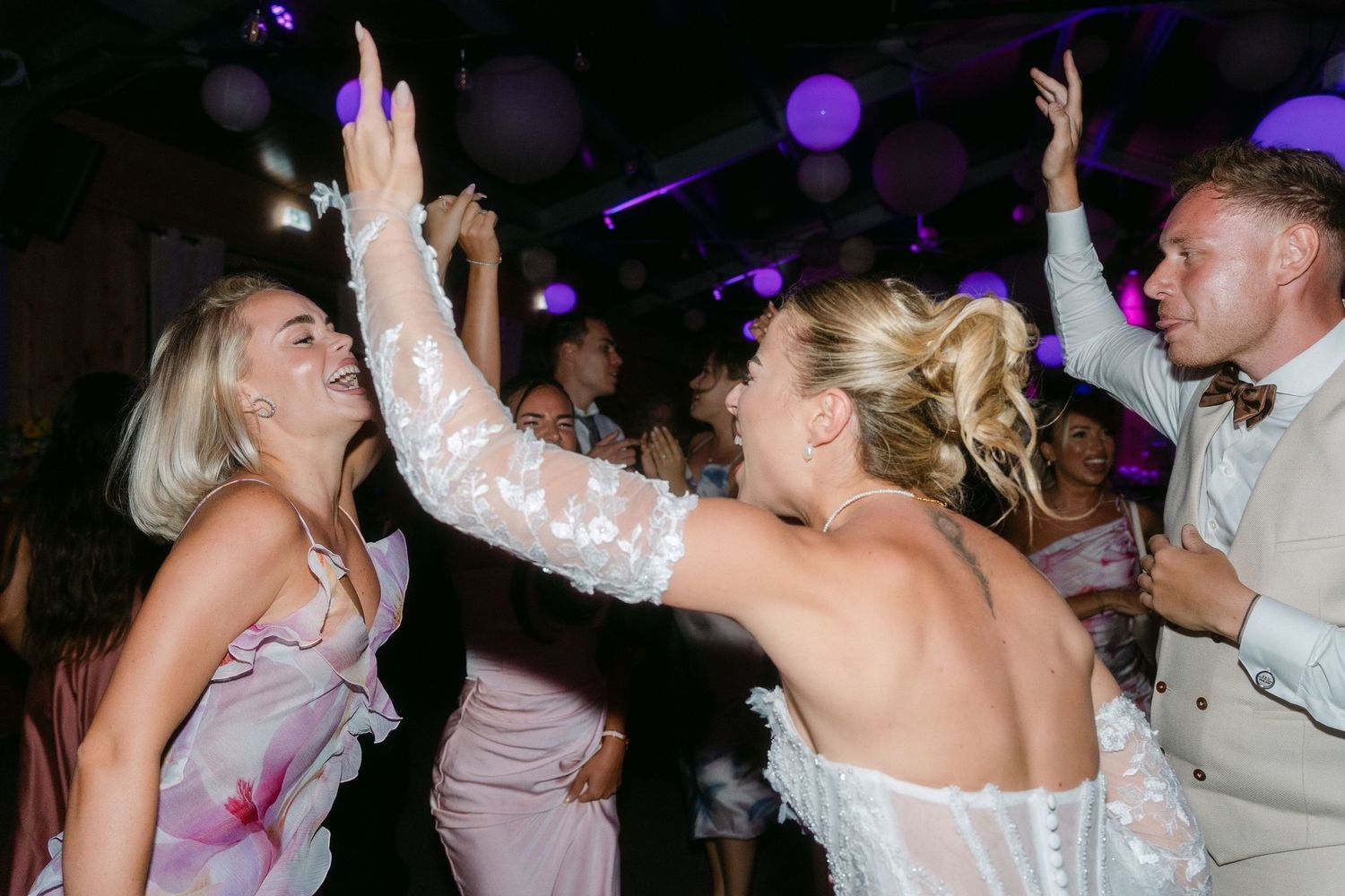 People dancing and celebrating on a dark dance floor with purple lighting at a wedding reception.