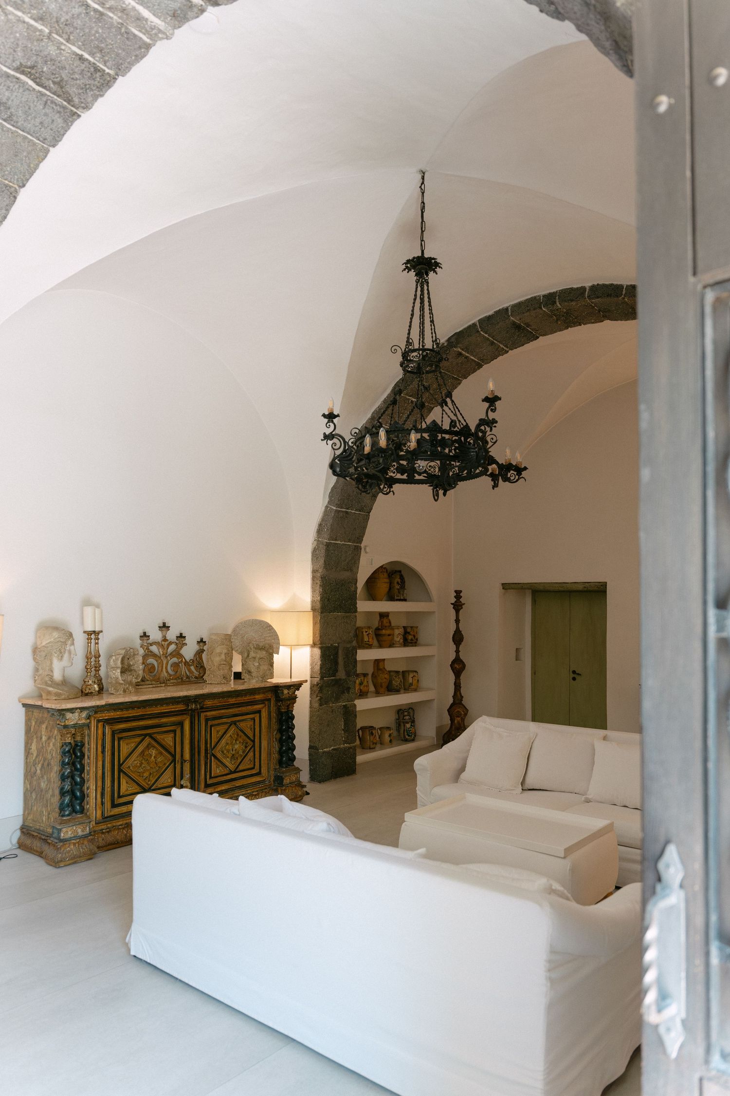 Elegant vaulted ceiling living room with white sofas, ornate chandelier, stone arch, and antique wooden sideboard with decorative objects.