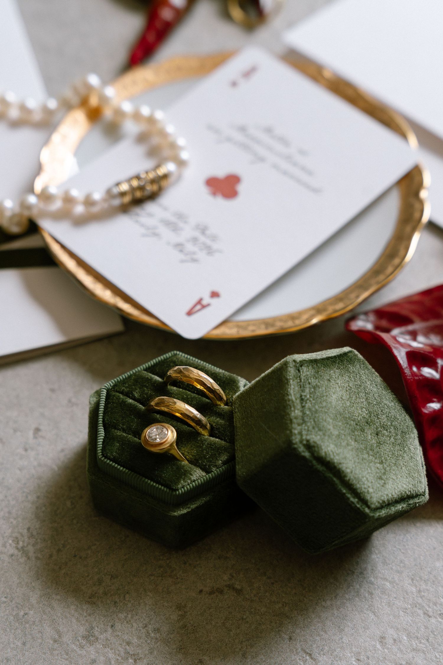 Gold wedding rings in green velvet hexagon box beside pearl bracelet and ace of hearts card on gold tray.