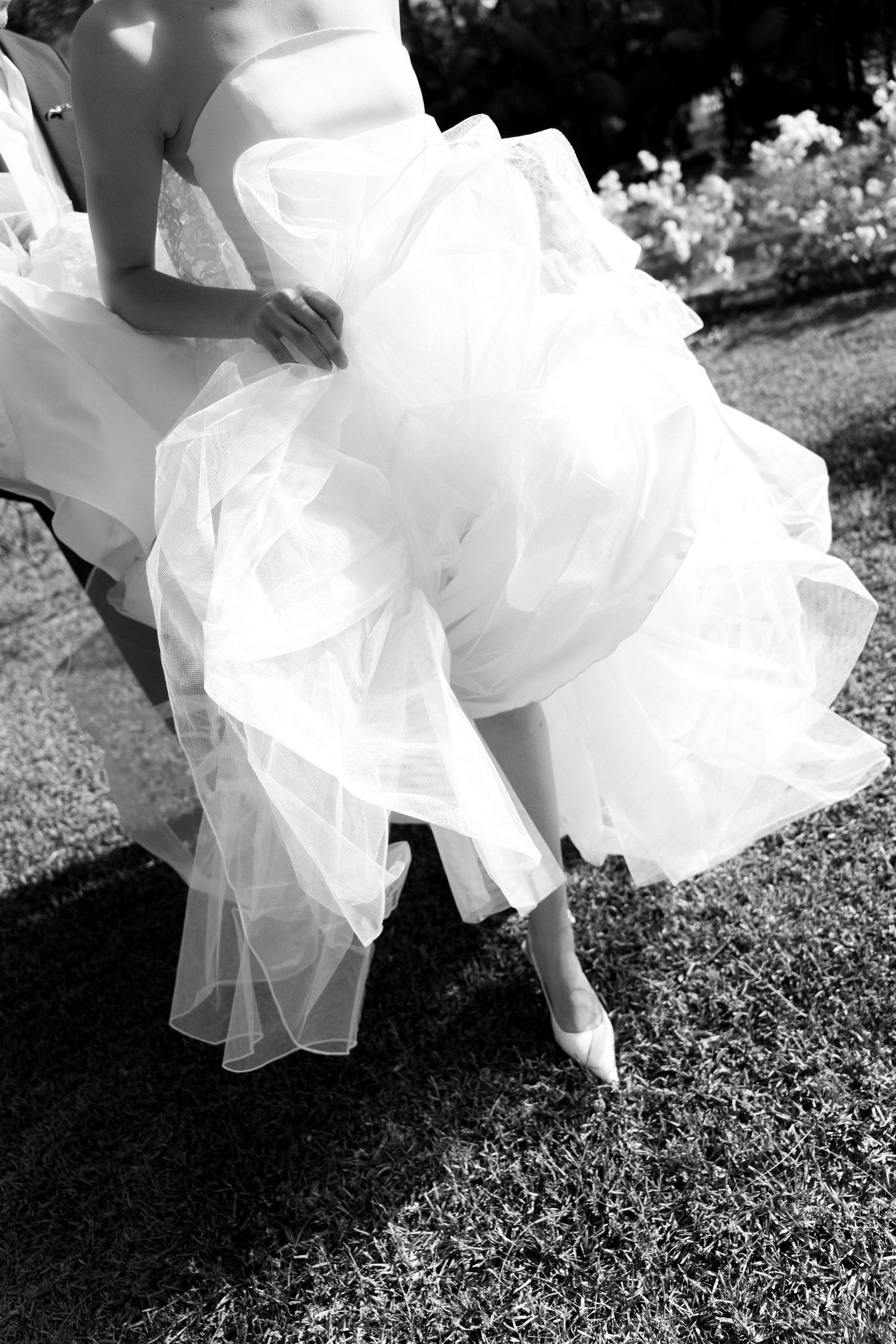 Black and white photo of a bride in a voluminous ruffled wedding gown and heels, seated outdoors on a sunny day.